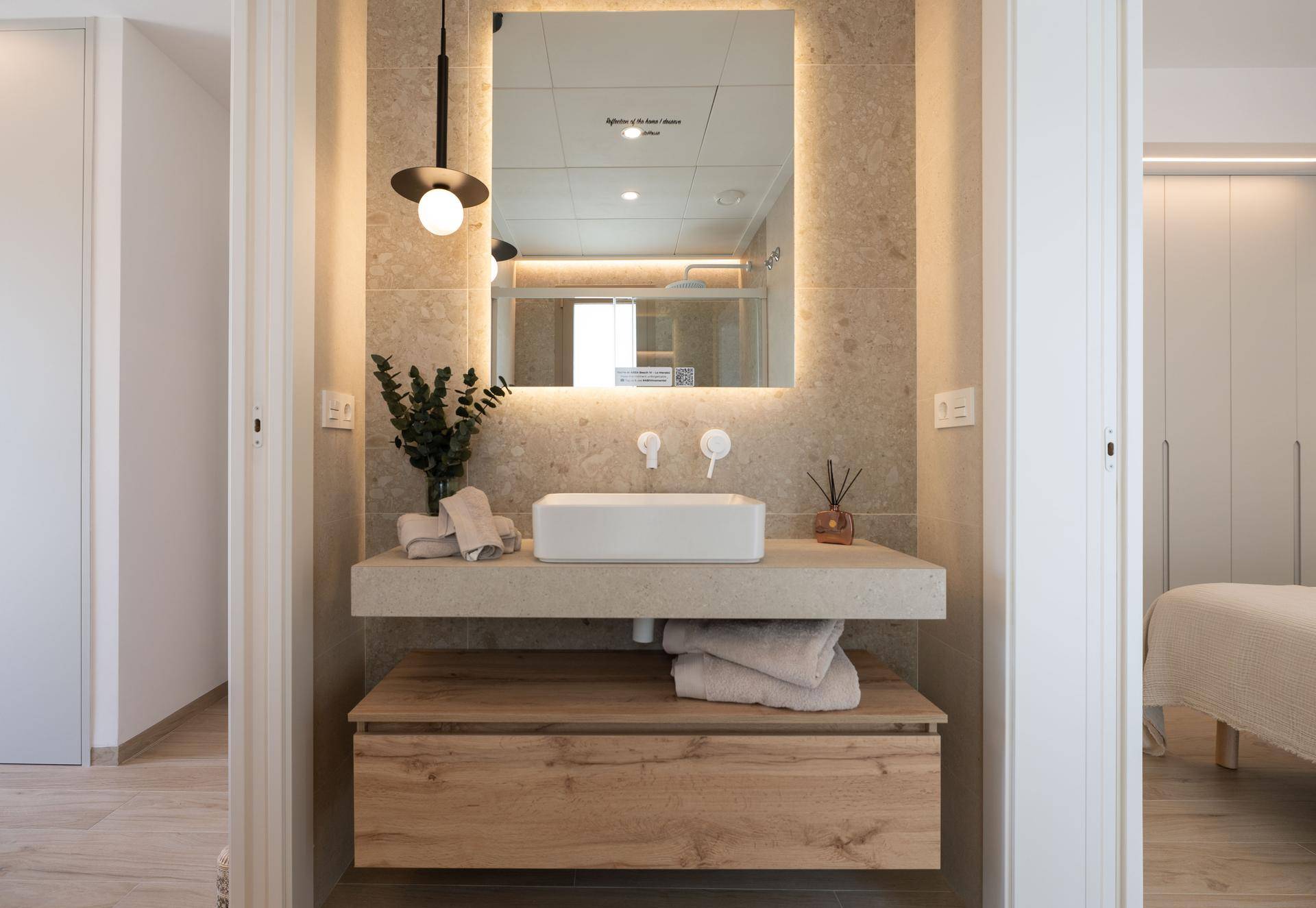 Modernes Badezimmer mit Einzelwaschbecken, beleuchtetem Spiegel und Zugang zum Schlafzimmer.