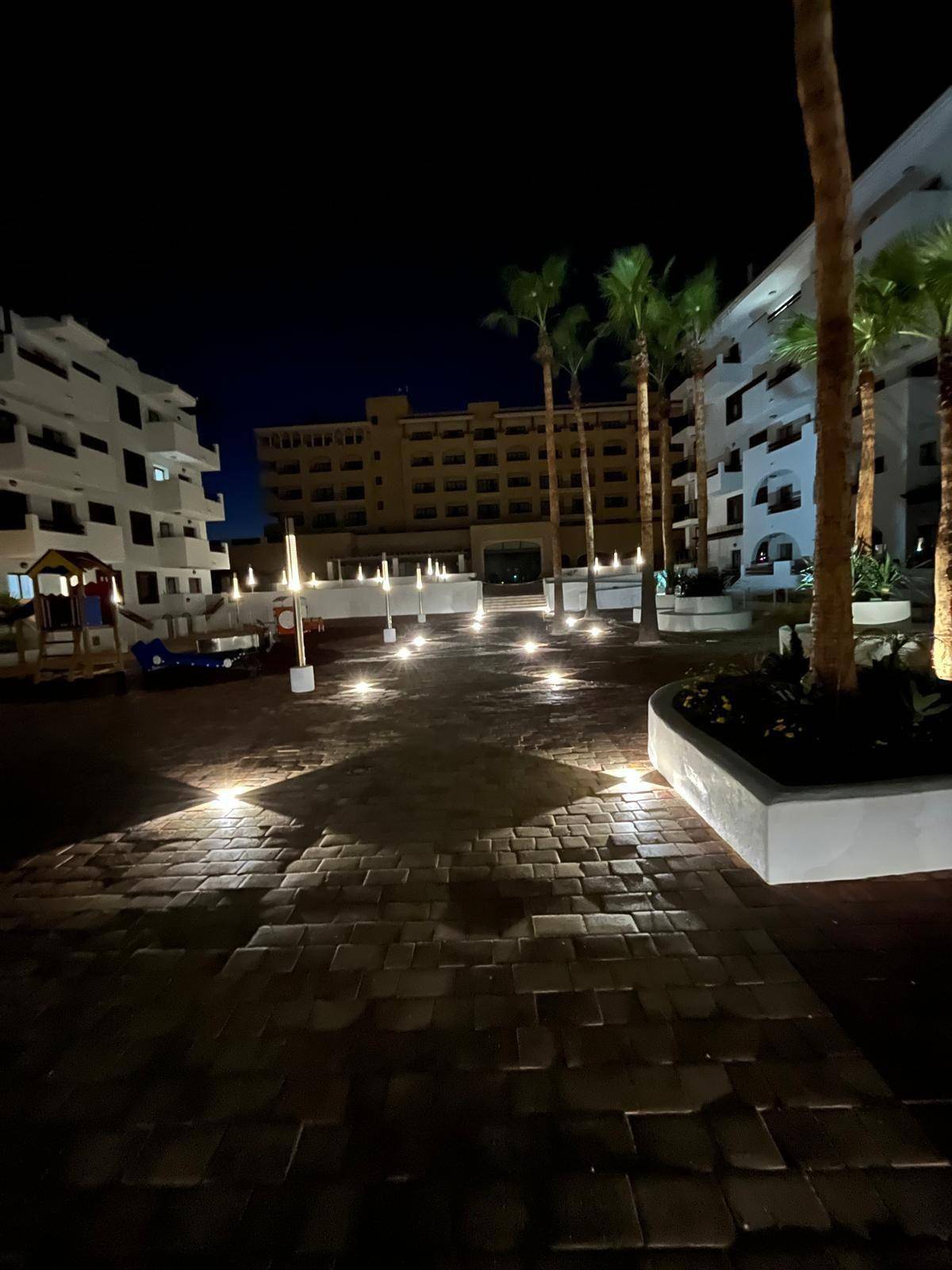 Beleuchteter Weg durch die Apartmentanlage bei Nacht mit Palmen und Gebäuden im Hintergrund.