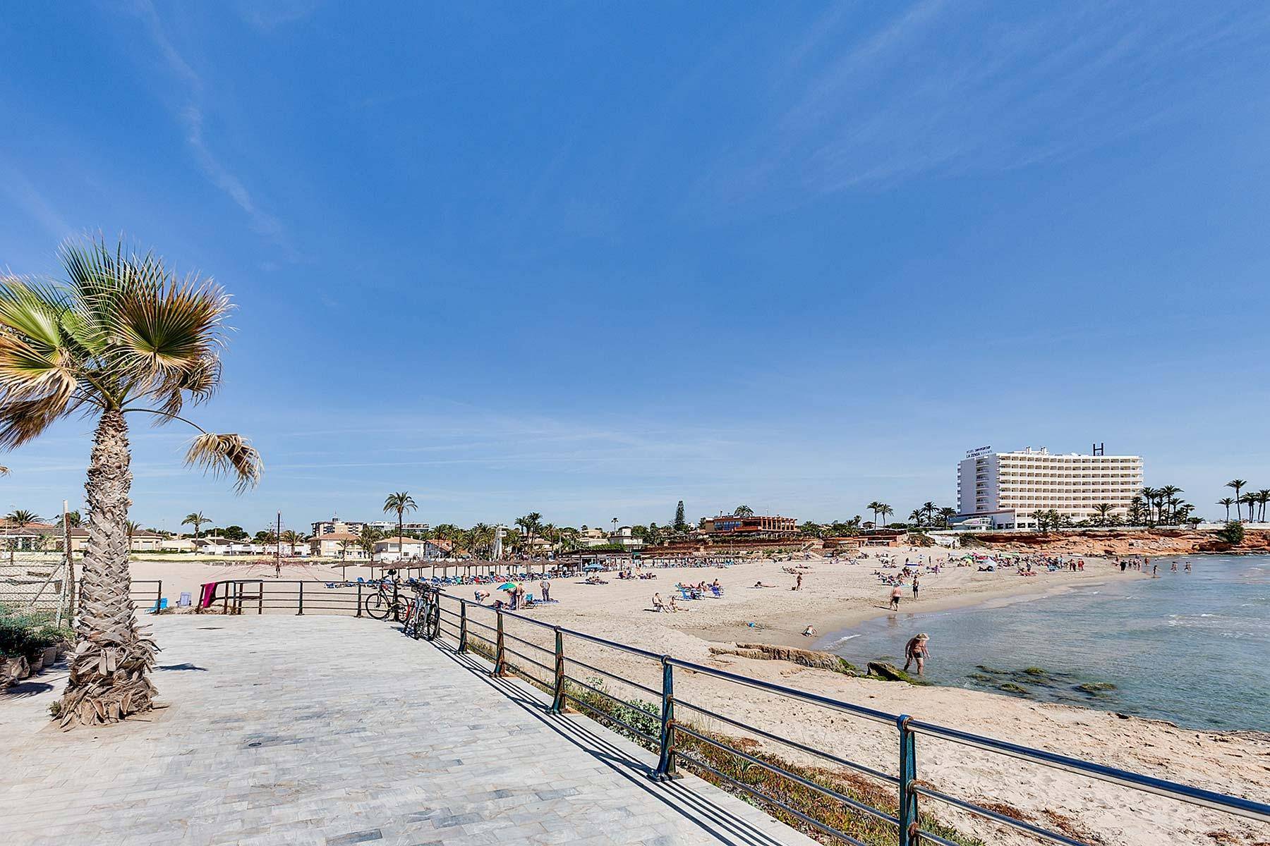 Promenade mit Blick auf einen belebten Sandstrand, das Meer und ein Hotelgebäude in der Ferne.