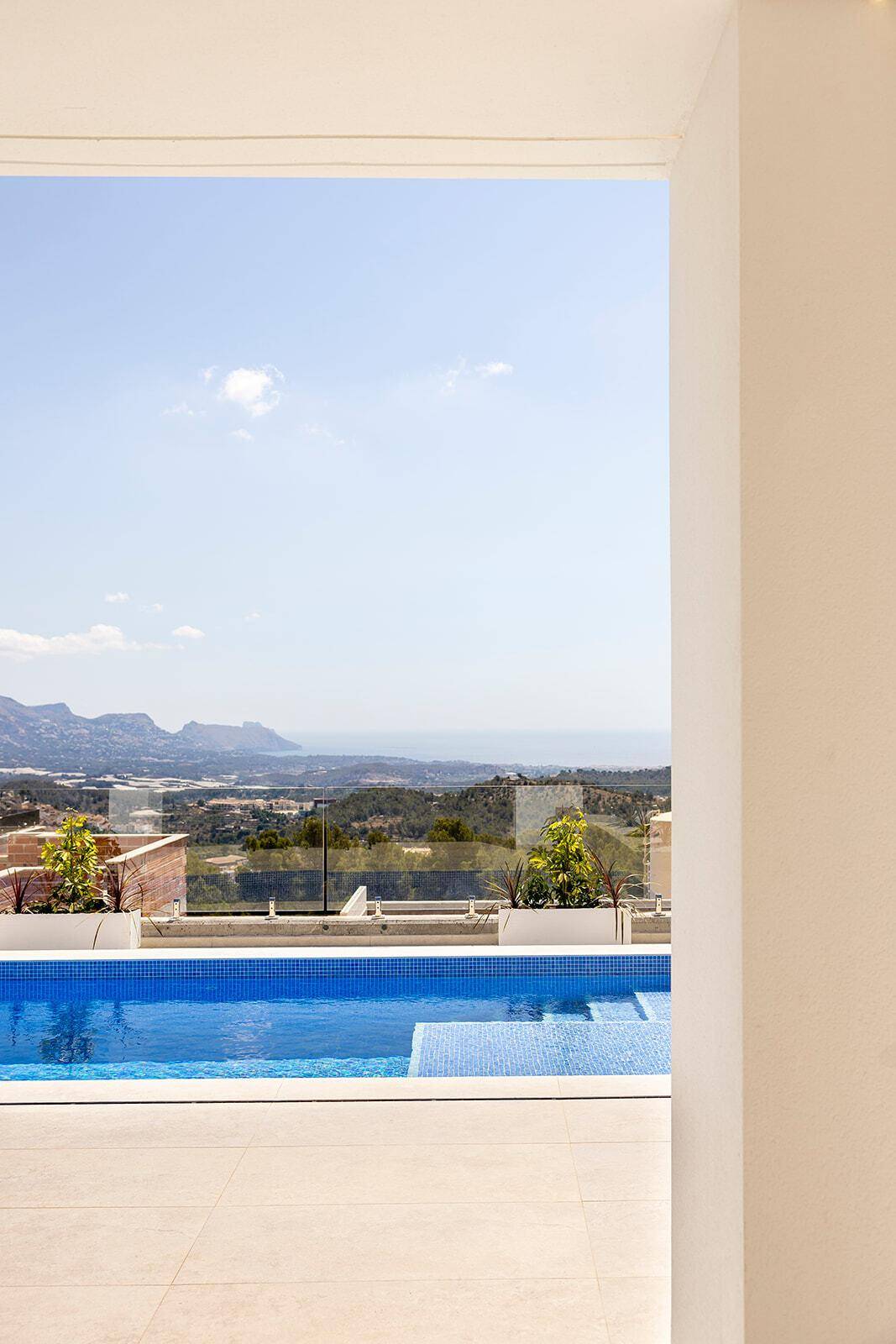 Blick von innen auf den Pool und die weite Landschaft mit Bergen und Meer am Horizont.