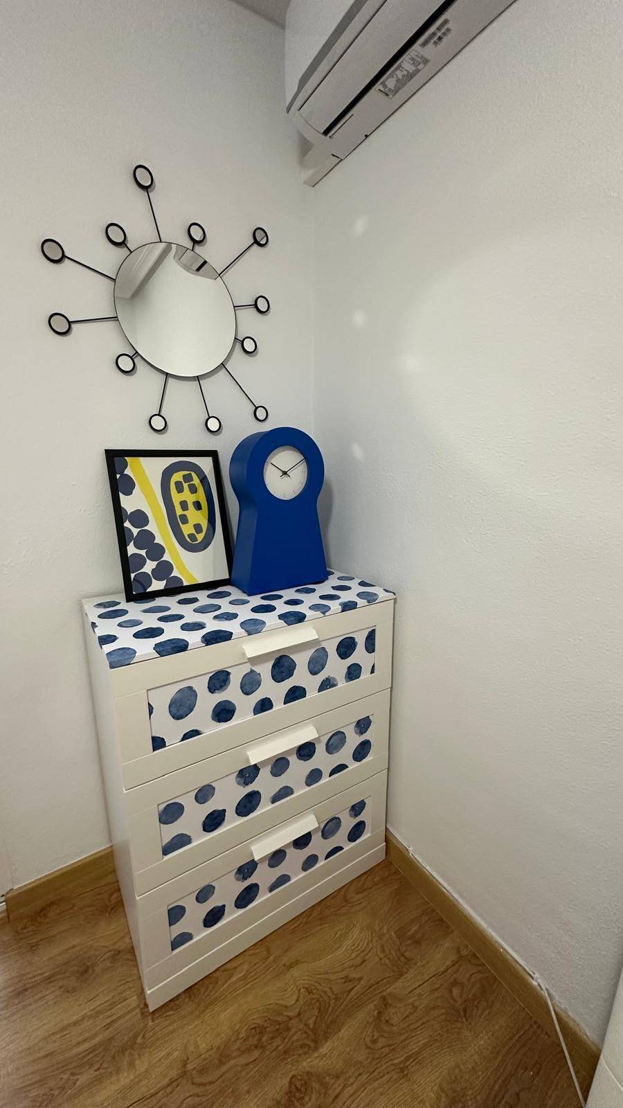 Nachttisch mit blauer Uhr und Spiegel, ergänzt durch eine Klimaanlage an der Wand.