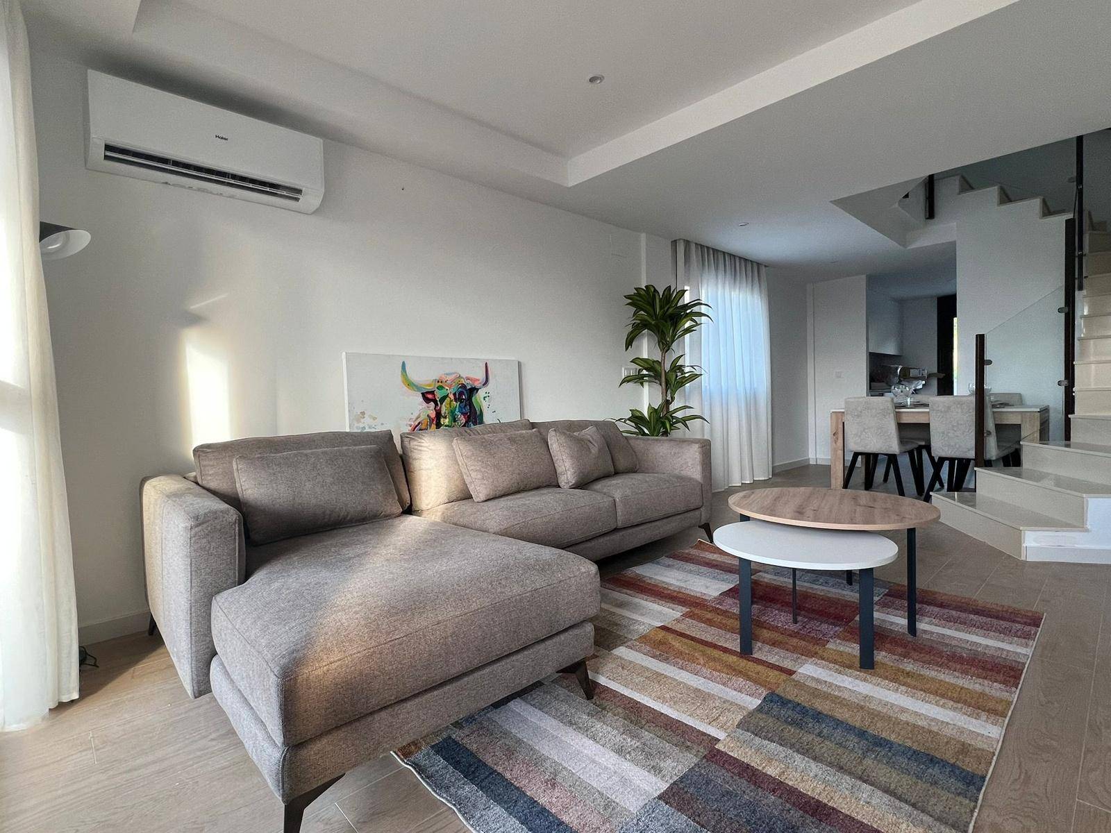 Heller Wohn- und Essbereich mit beigem Sofa, farbigem Teppich und Treppe.