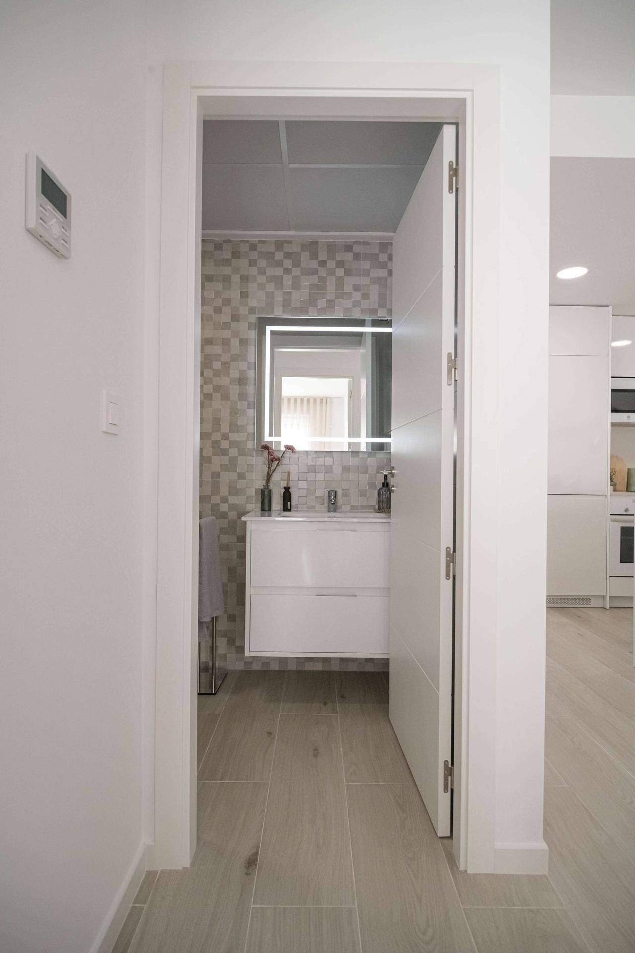Heller Flur mit Blick in ein modernes Badezimmer mit beleuchtetem Spiegel und Waschtisch.