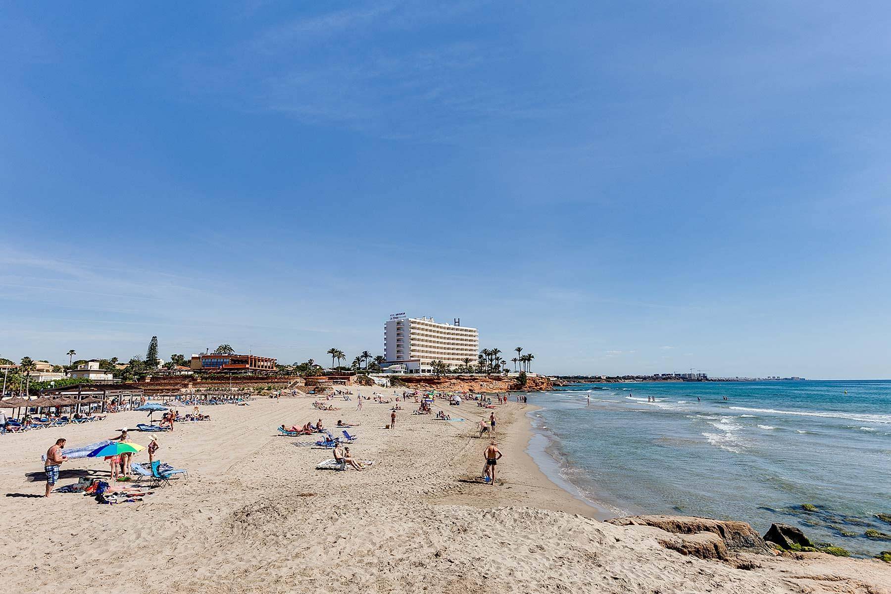 Belebtes Strandpanorama mit Badegästen, Sonnenschirmen und einem Hotelgebäude am Horizont.