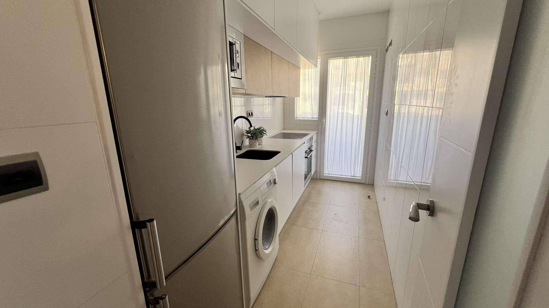 Helle Küche mit Kühlschrank, Waschmaschine, Spüle und Fenster, in Weiß und Holz gehalten.