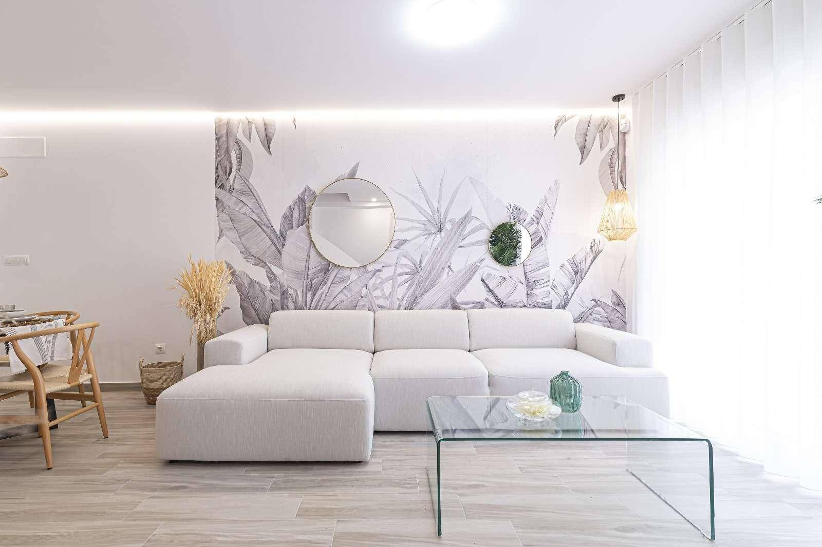 Heller Wohnbereich mit großem Ecksofa, Glastisch und tropischer Wanddekoration.
