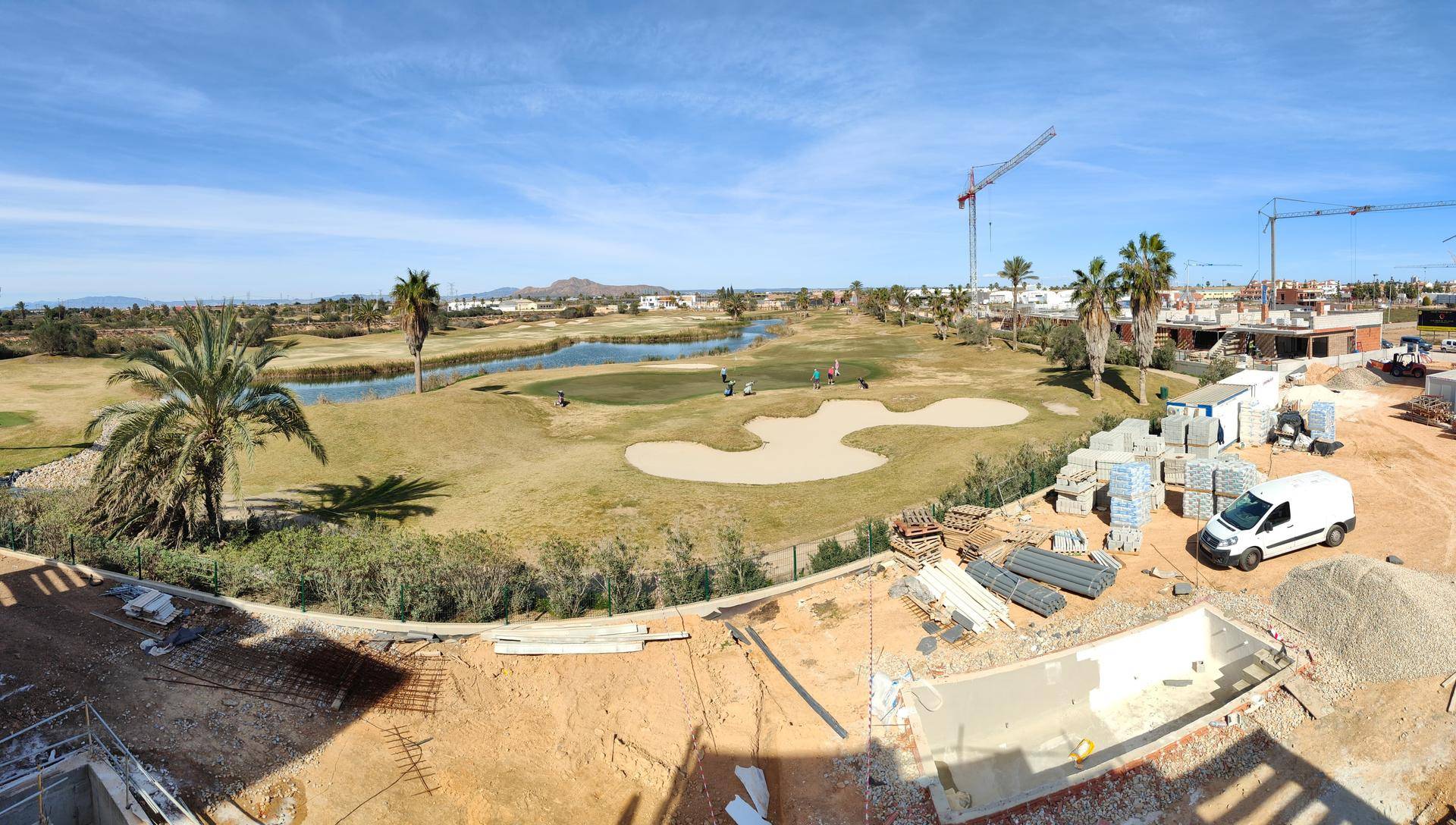 Baustelle neben einem Golfplatz mit Kränen und Materiallager unter blauem Himmel.