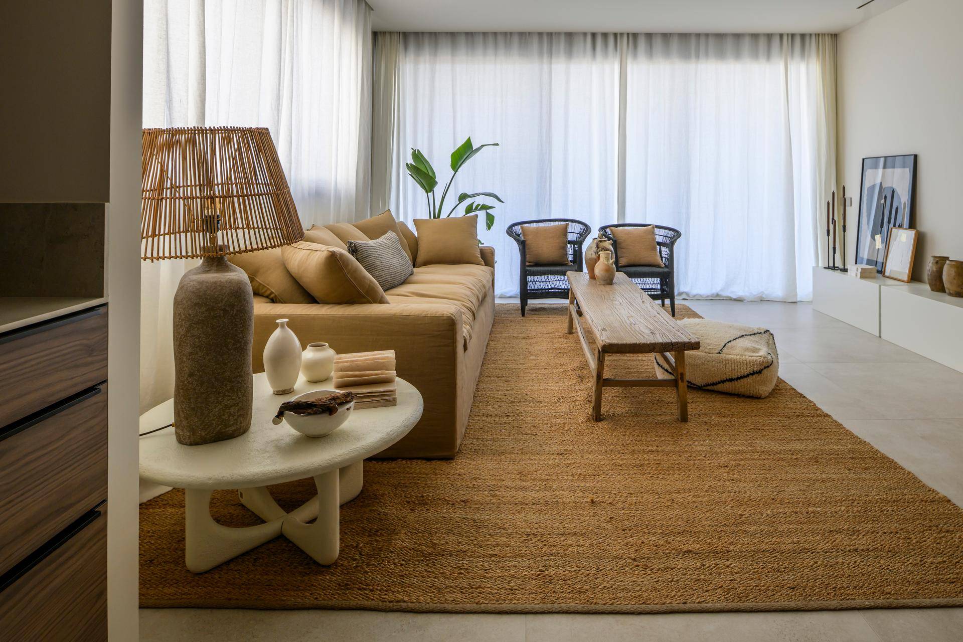Geräumiger Wohnbereich mit beigem Sofa, Holztisch und großer Pflanze, umgeben von hellen Vorhängen.