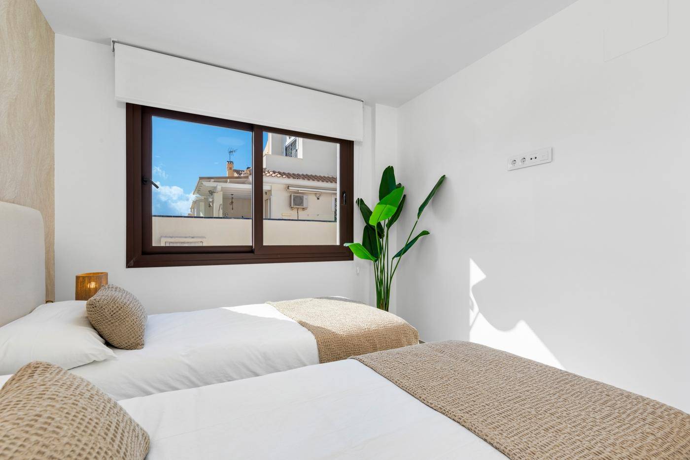 Helles Schlafzimmer mit zwei Einzelbetten, einem Fenster und einer grünen Pflanze als Dekoration.