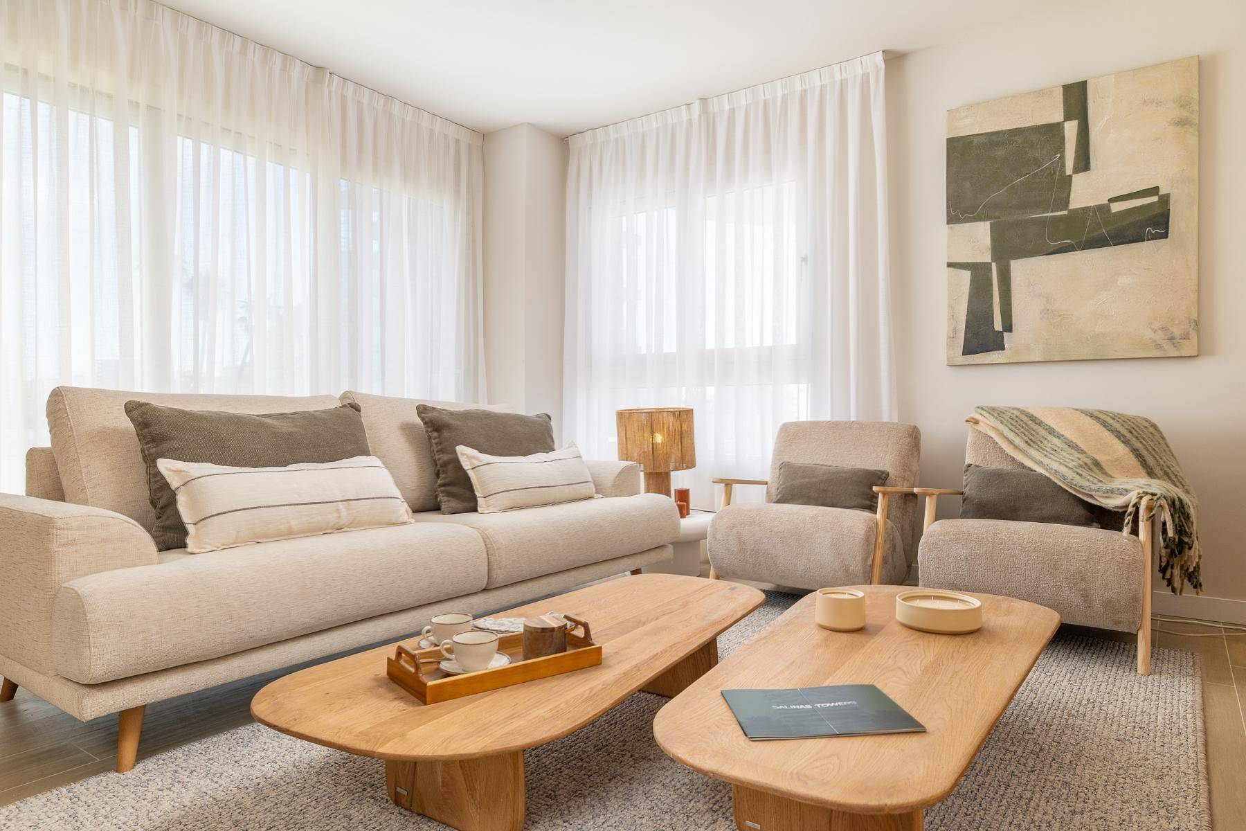 Gemütliches Wohnzimmer mit beigem Sofa, zwei Sesseln und eleganten Holztischen vor großen Fenstern.