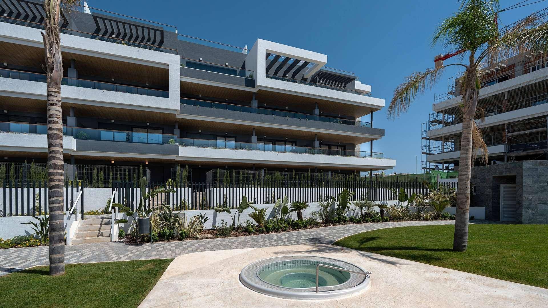Modernes Apartmentgebäude mit Whirlpool im Gartenbereich und Palmen unter strahlendem Sonnenschein.