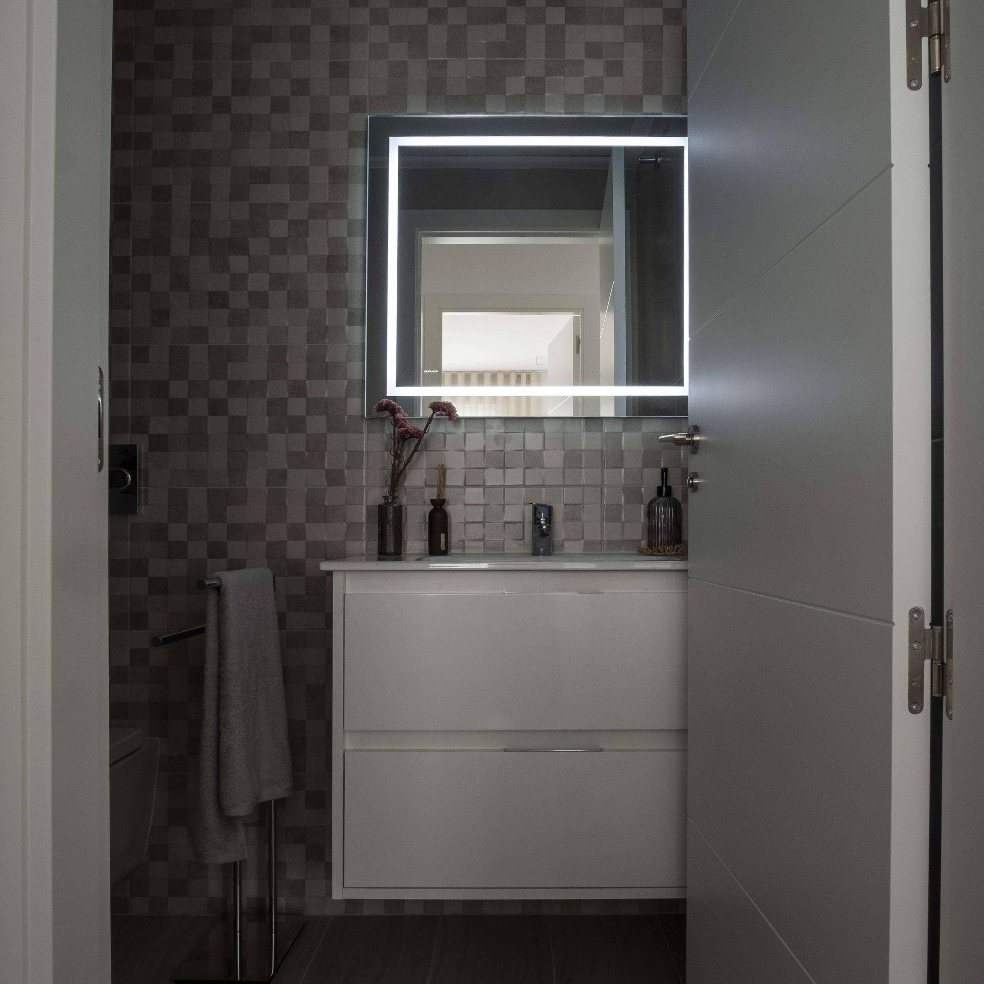 Modernes Badezimmer mit Waschtisch, beleuchtetem Spiegel und grauen Mosaikfliesen.