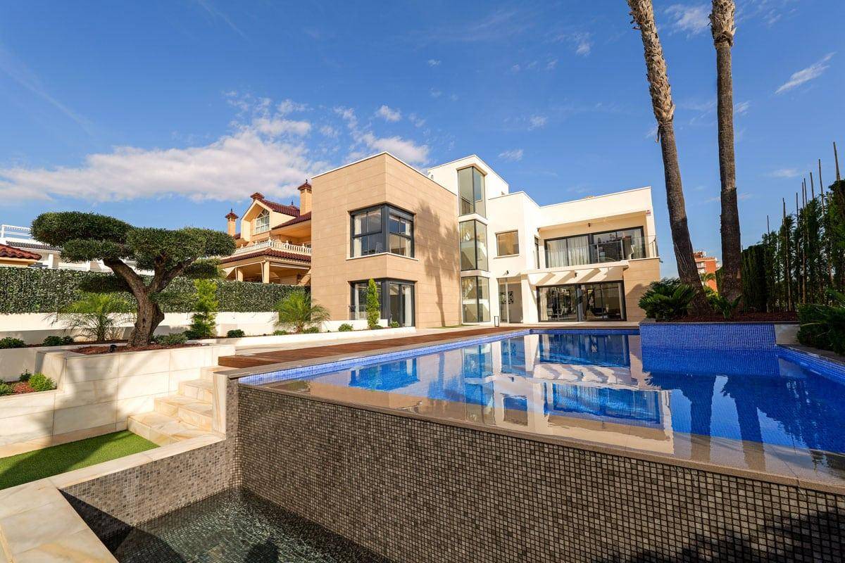 Luxuriöse Villa mit großem, mehrstuigem Pool und gepflegter Gartenanlage unter strahlendem Himmel.