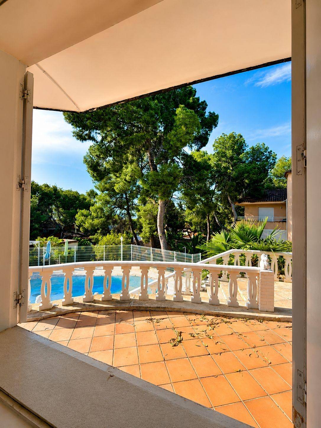 Blick aus einem Fenster auf die Poolterrasse und die grüne Umgebung unter strahlend blauem Himmel.