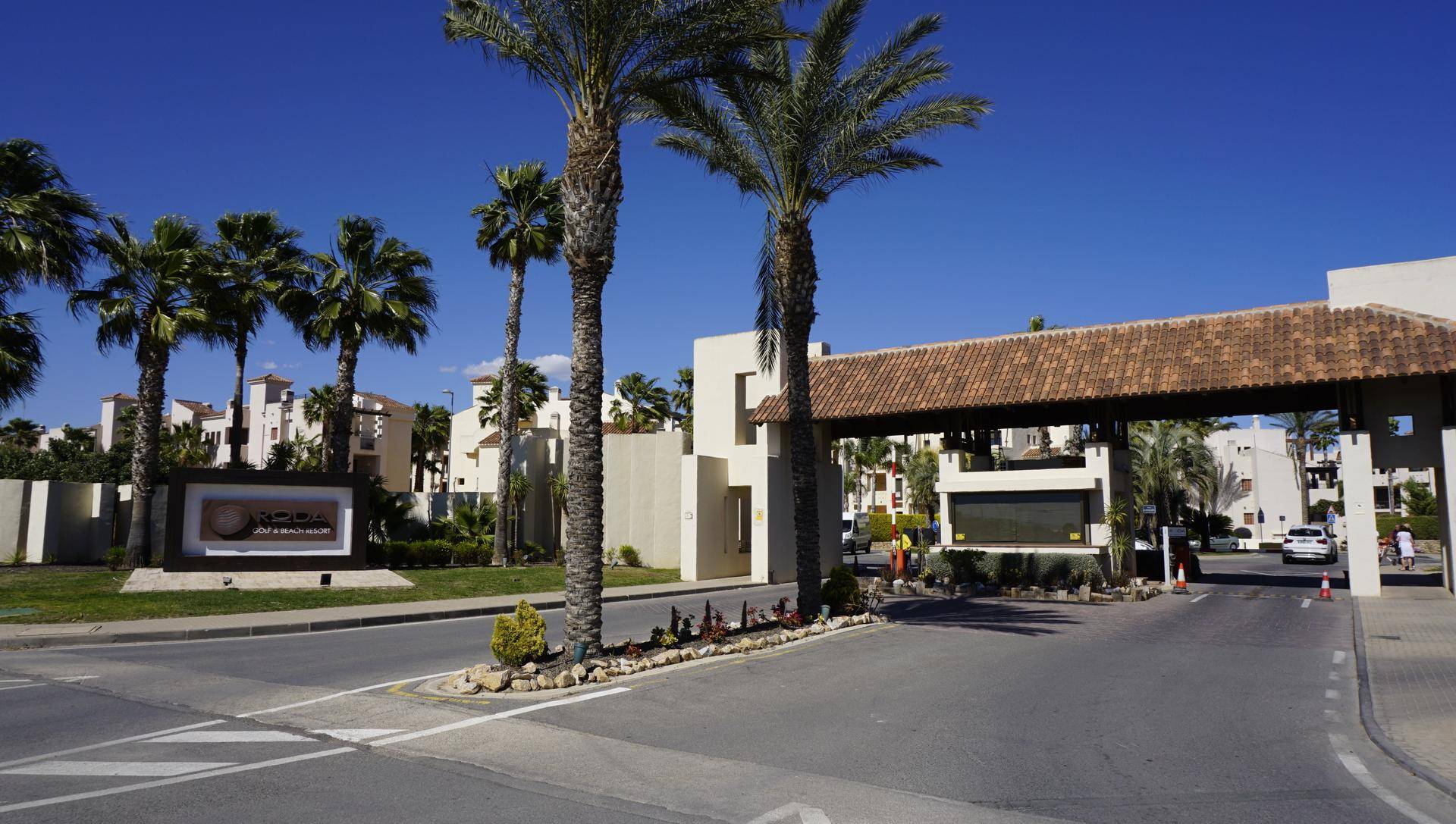 Eingangsbereich des Resorts mit Tor, Palmen und Zufahrtsstraße unter blauem Himmel.