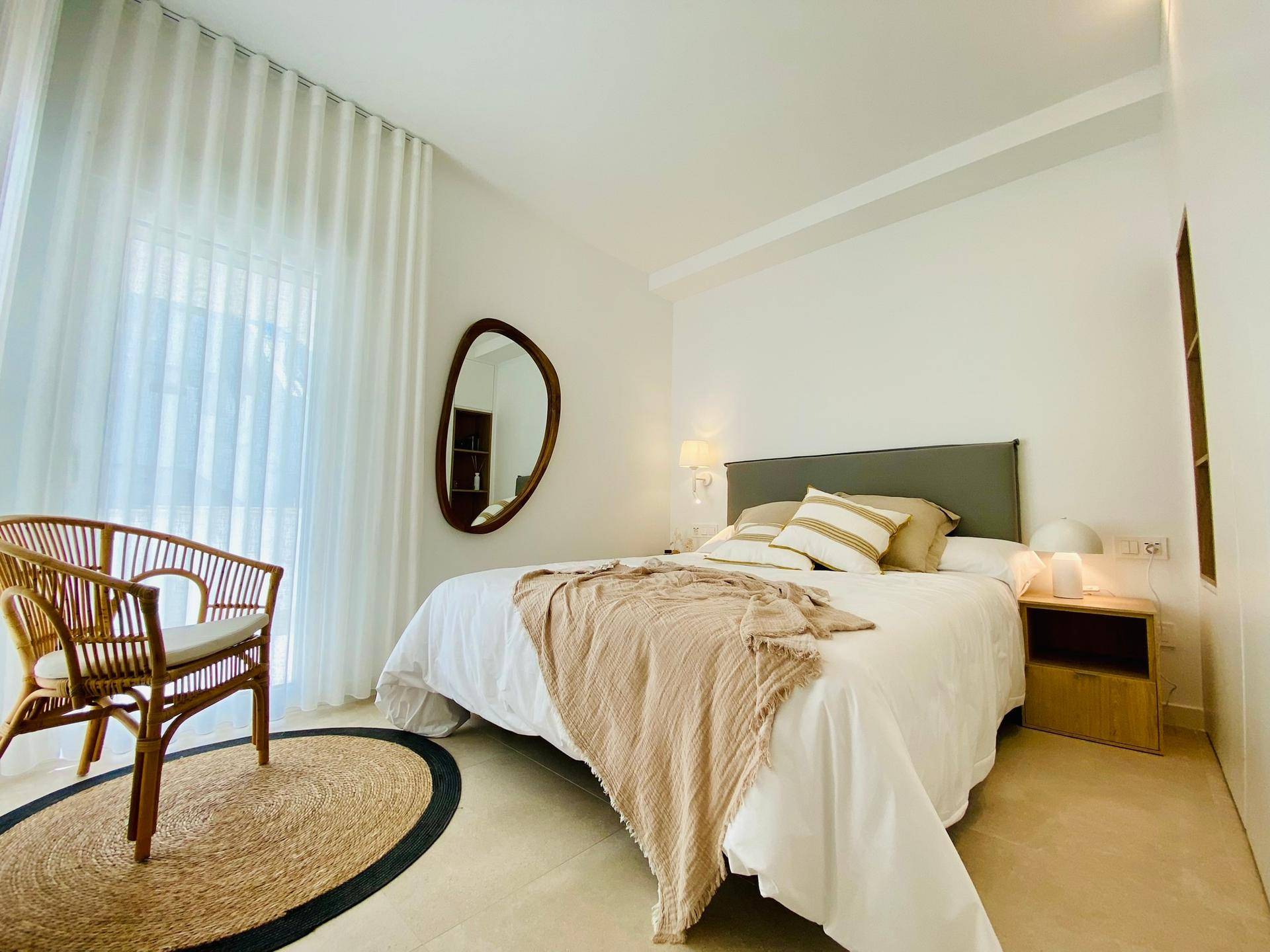 Gemütliches Schlafzimmer mit großem Fenster, Rattan-Sessel und heller Einrichtung.