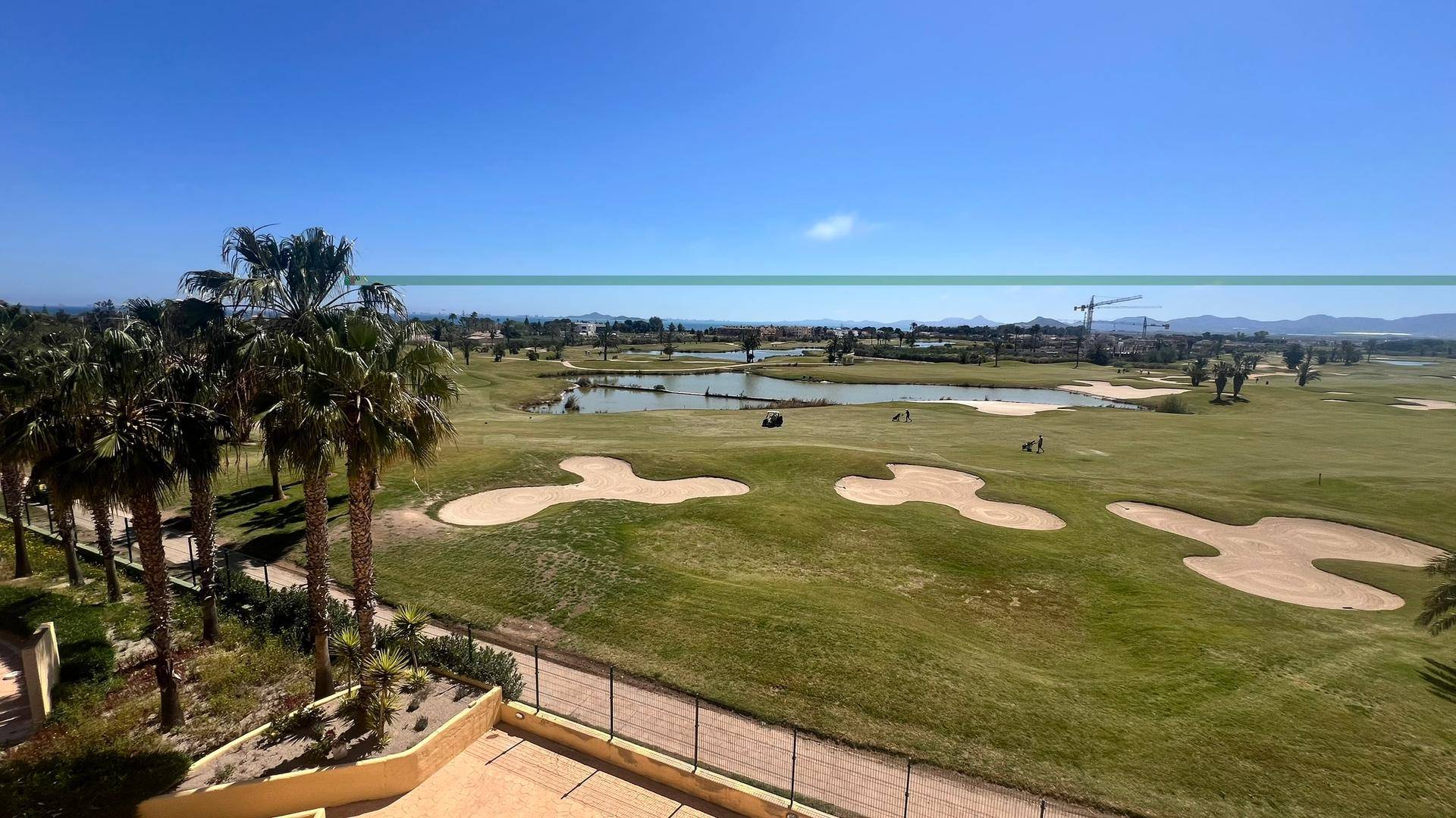 Weitläufiger Blick auf einen grünen Golfplatz mit Wasserhindernissen und Palmen unter blauem Himmel.