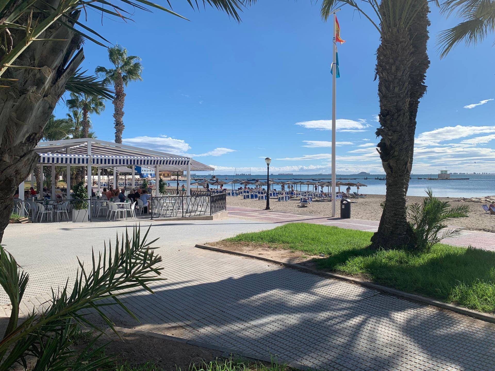 Belebter Sandstrand mit einem Strandcafé und vielen Sonnenliegen unter blauem Himmel.