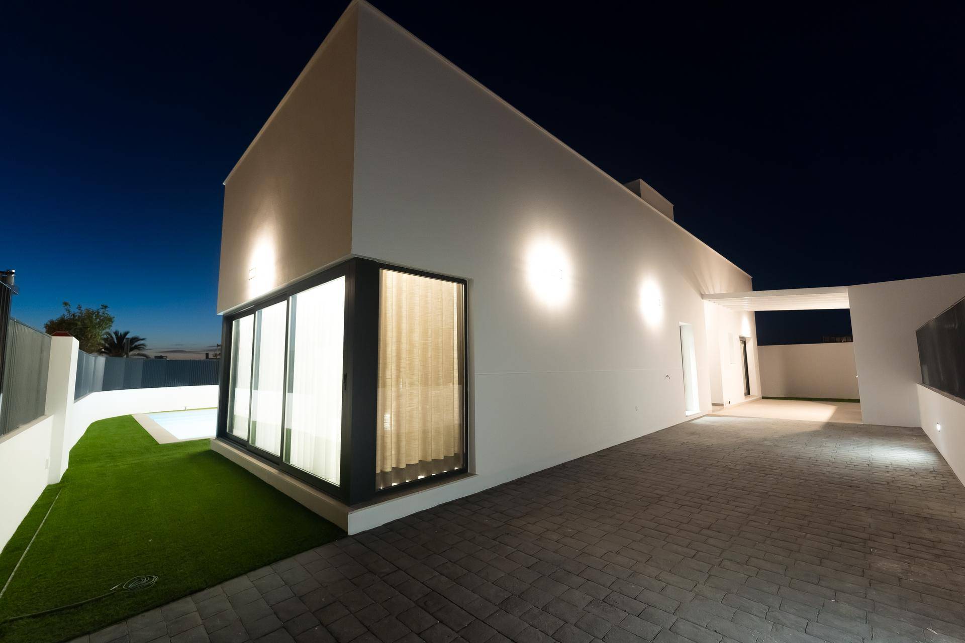 Moderne Hausfassade bei Nacht mit beleuchteter Auffahrt und großen Fenstern.