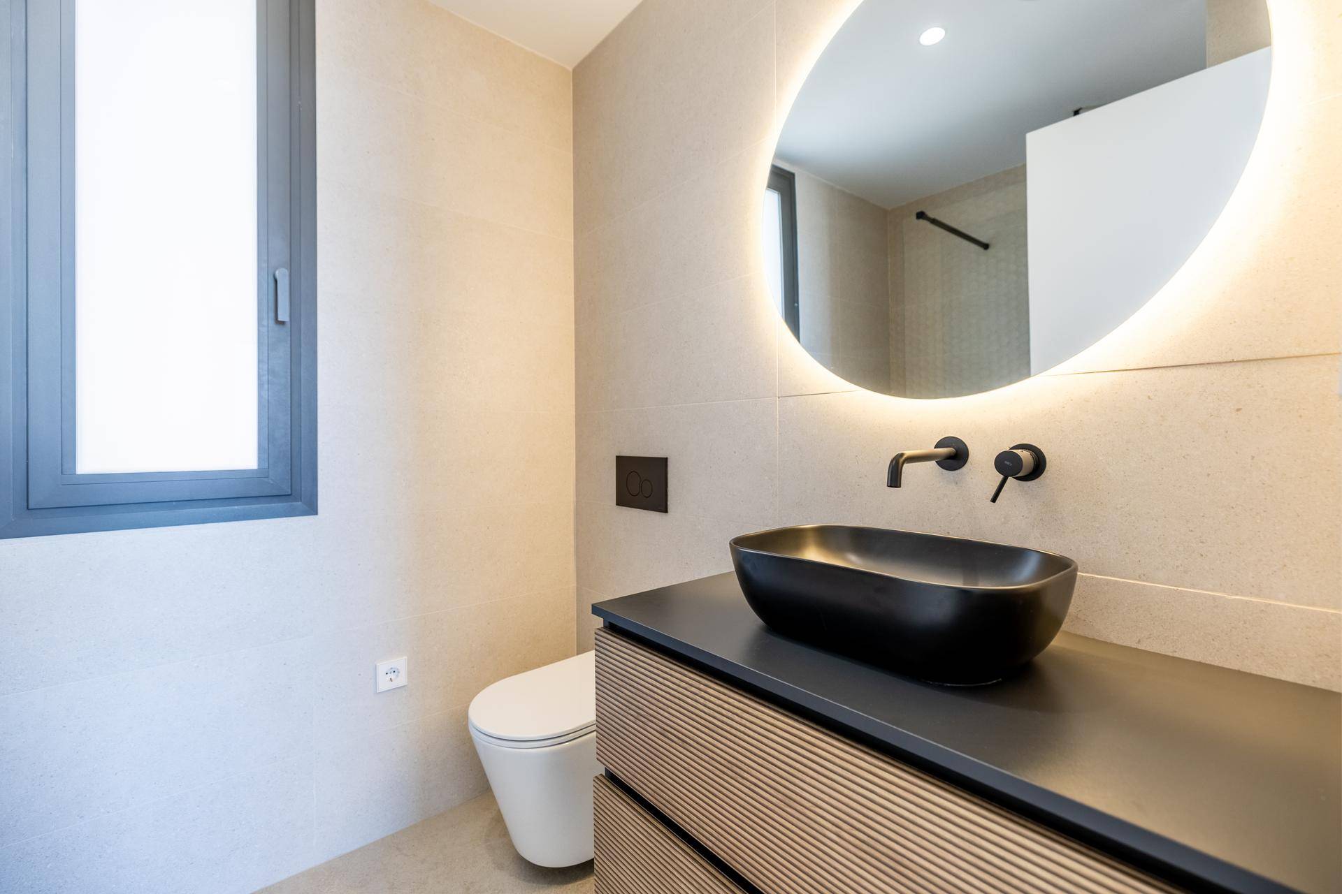 Stilvolles Badezimmer mit rundem beleuchtetem Spiegel, schwarzem Waschbecken und Fenster.