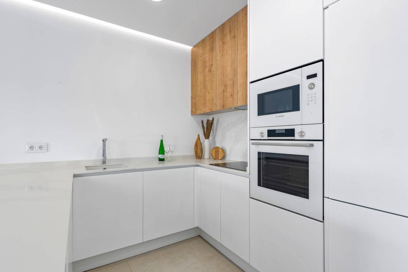 Moderne weiße Küche mit Holzdetails, integrierten Geräten und heller Arbeitsplatte.