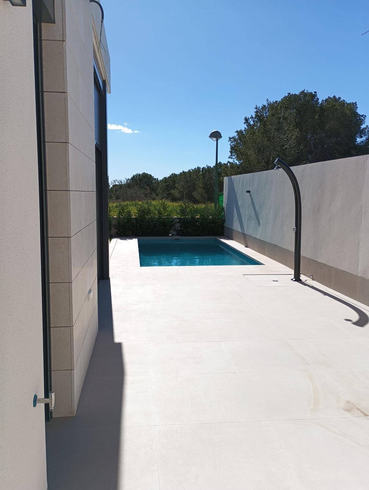 Schmaler Poolbereich mit gefliester Terrasse und Außendusche, umgeben von einer hohen Mauer.