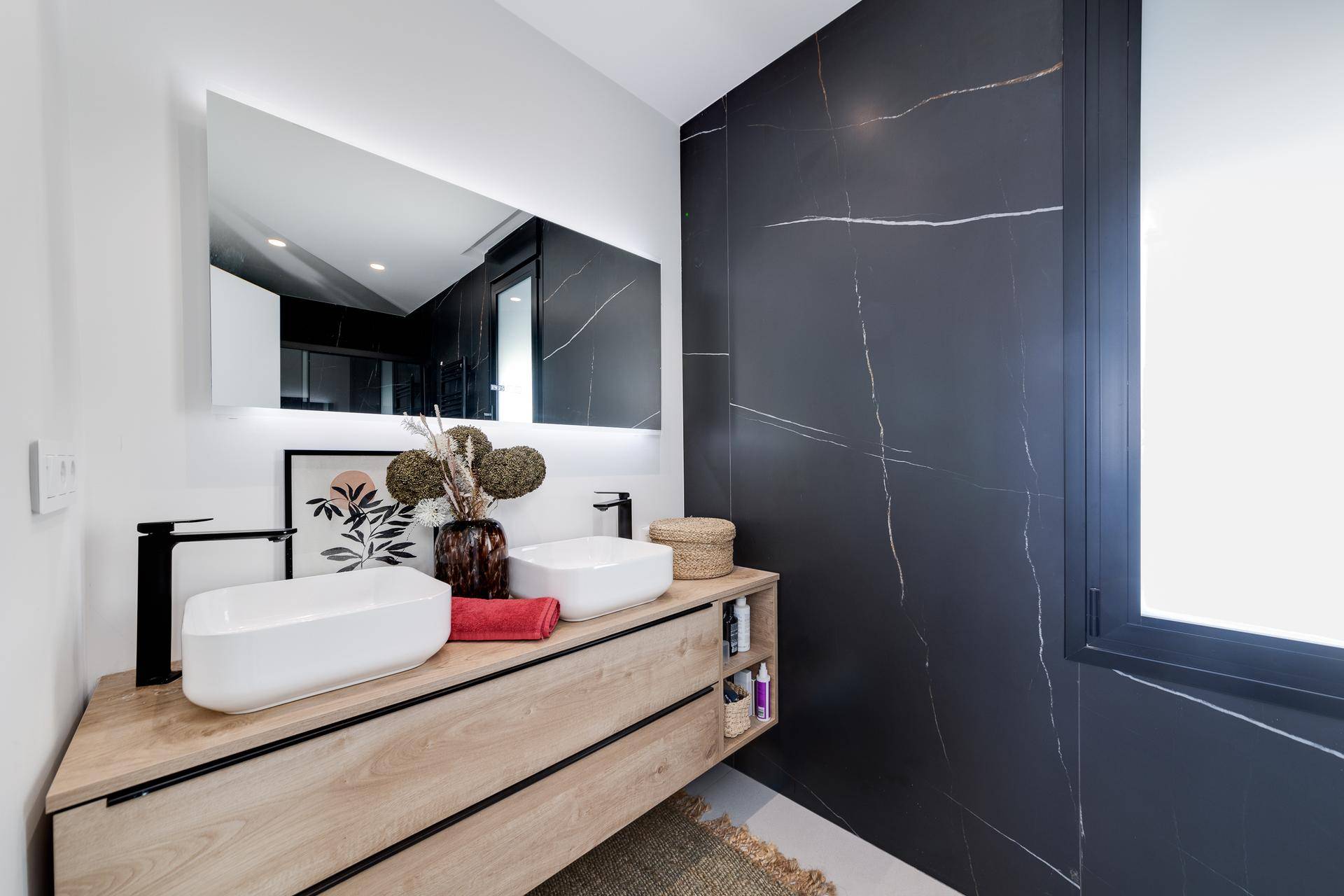 Elegantes Badezimmer mit Doppelwaschbecken, Holzunterschrank und dunkler Marmorwand, beleuchtetem Spiegel.