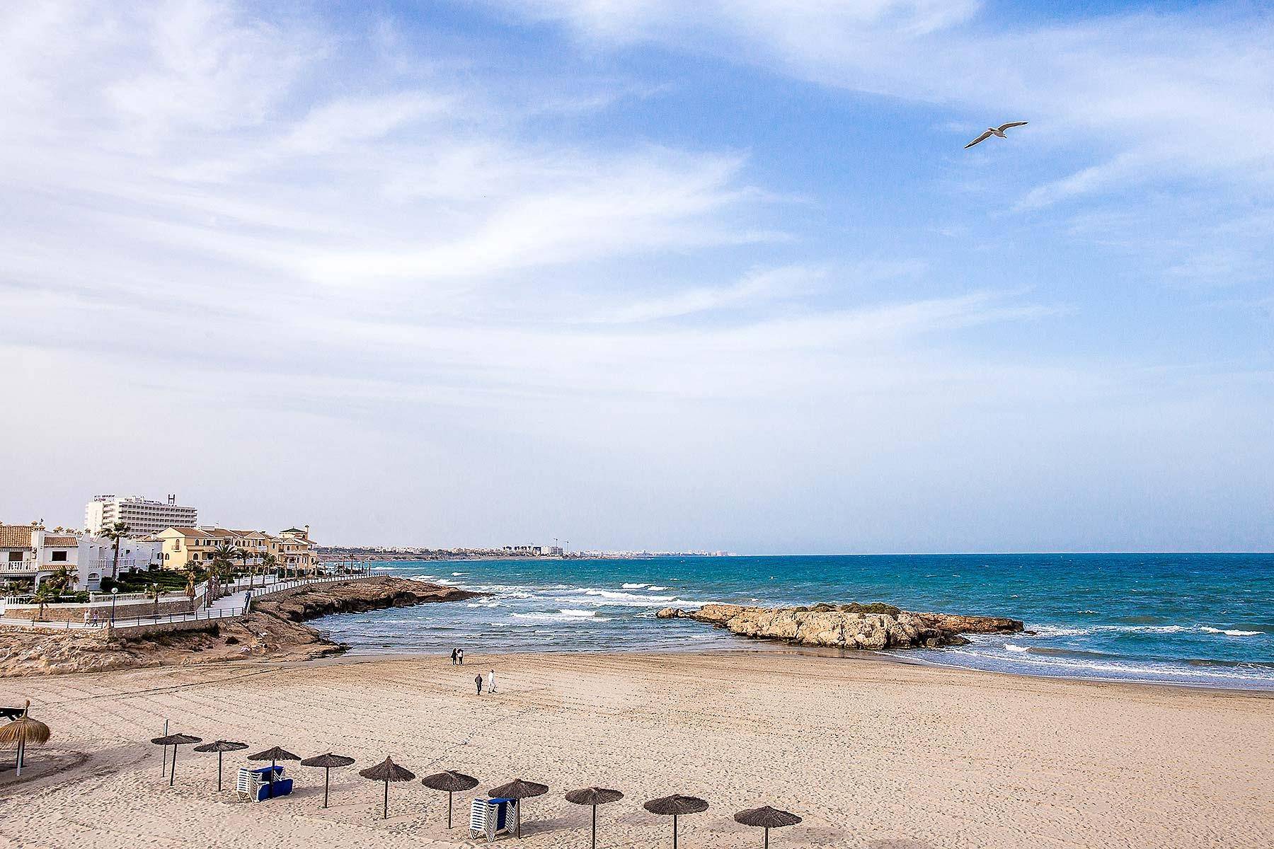 Ruhiger Strandabschnitt mit wenigen Sonnenschirmen, Meerblick und Gebäuden am Horizont.