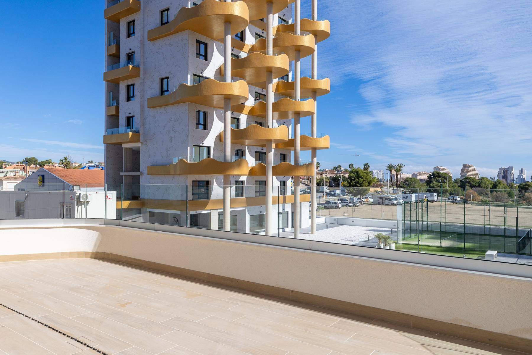 Geräumige Terrasse mit Glasbrüstung, Blick auf ein modernes Gebäude und Sportanlagen.