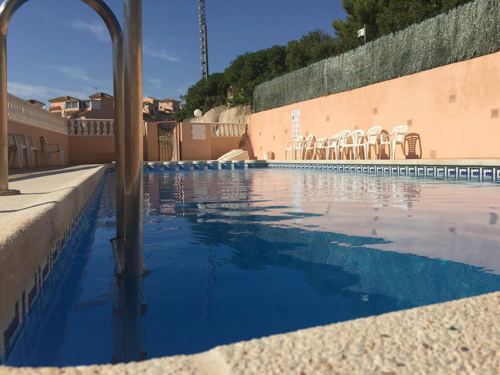 Gemeinschaftspool mit blauen Fliesen und umliegender Terrasse unter strahlendem Sonnenschein.