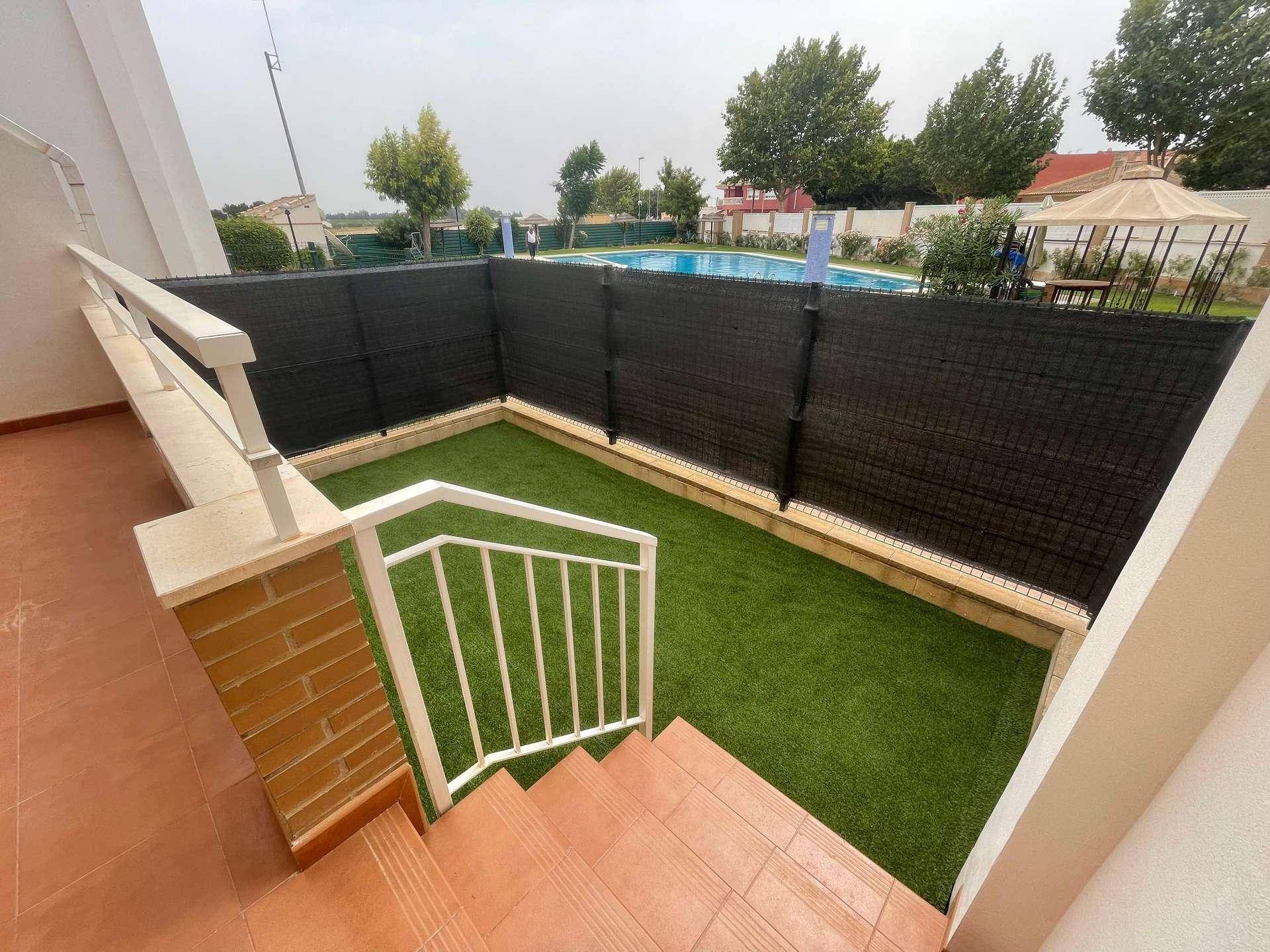Blick von der Terrasse auf den kleinen Gartenbereich mit Kunstrasen und den Gemeinschaftspool im Hintergrund.