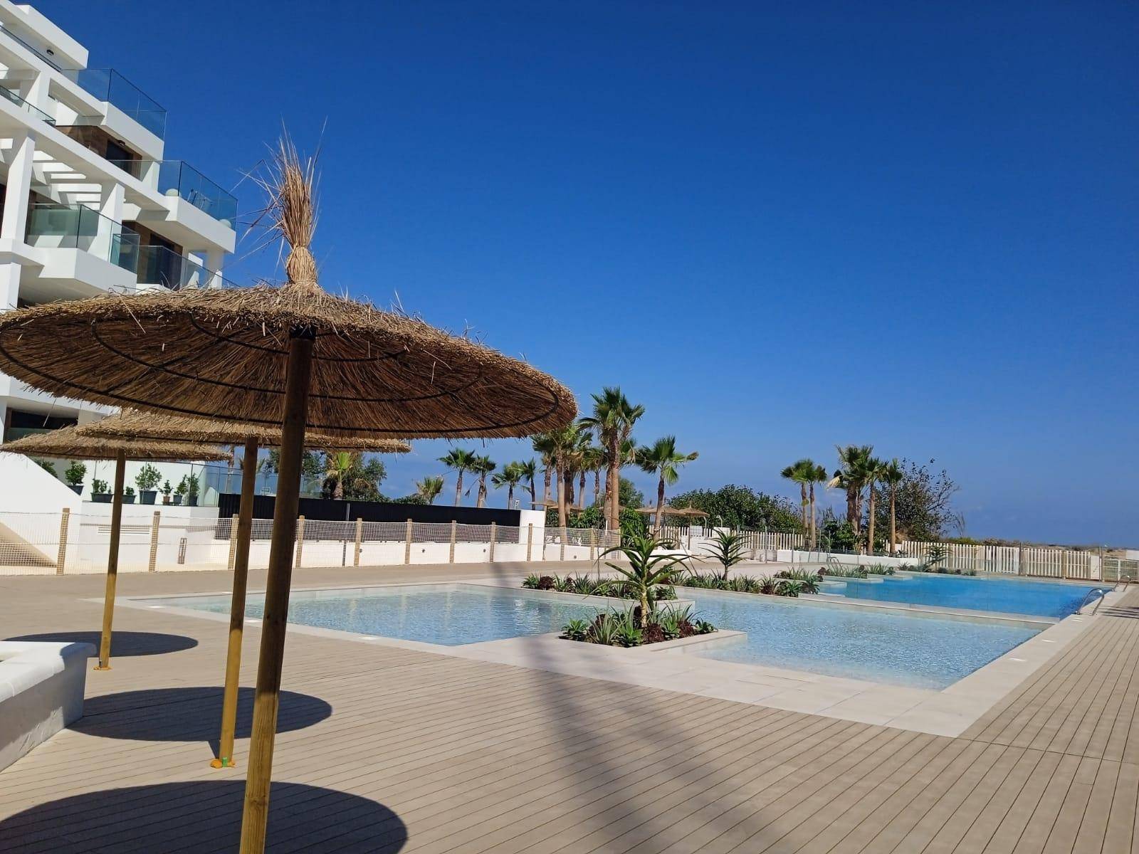 Weitläufiger Poolbereich mit Strohschirmen, Palmen und Blick auf das Meer in der Ferne.