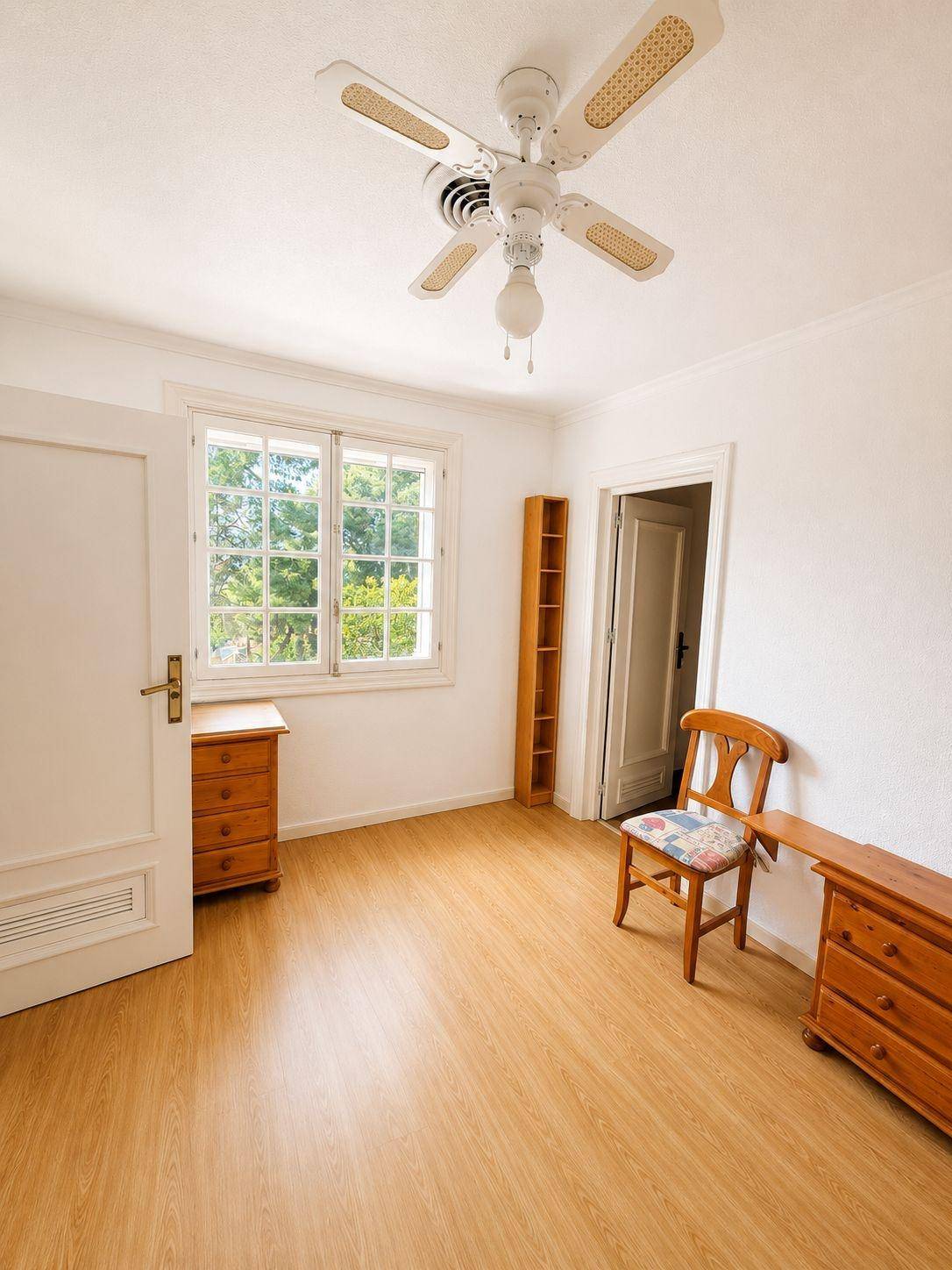 Gemütliches Zimmer mit Holzboden, Deckenventilator und einem Fenster mit Blick ins Grüne.