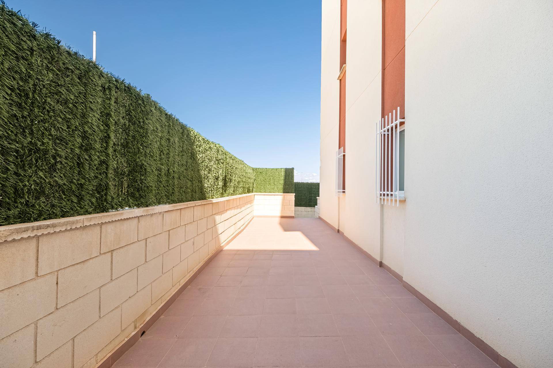 Lange Außenterrasse entlang der Gebäudefassade mit viel Sonnenlicht und Hecke.