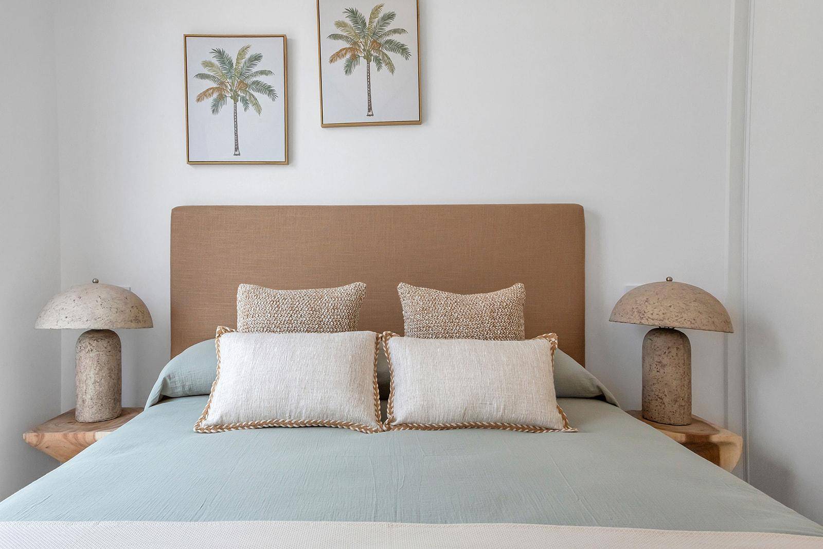 Helles Schlafzimmer mit beigem Kopfteil, zwei Nachttischlampen und Palmenbildern.