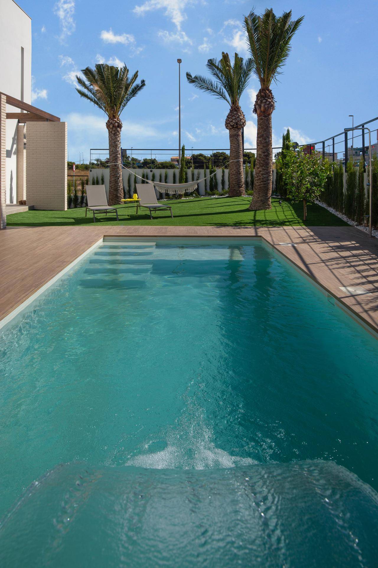 Nahaufnahme des Pools mit Stufen, umgeben von Holzdeck und Kunstrasen mit Palmen und Hängematte.