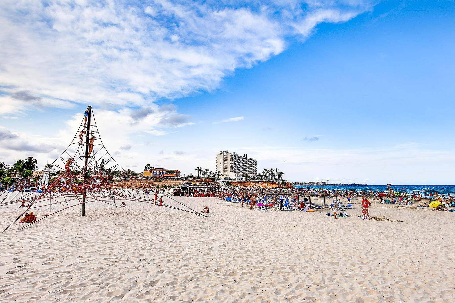 Breiter Sandstrand mit großem Klettergerüst, vielen Badegästen und einem Hotel im Hintergrund.