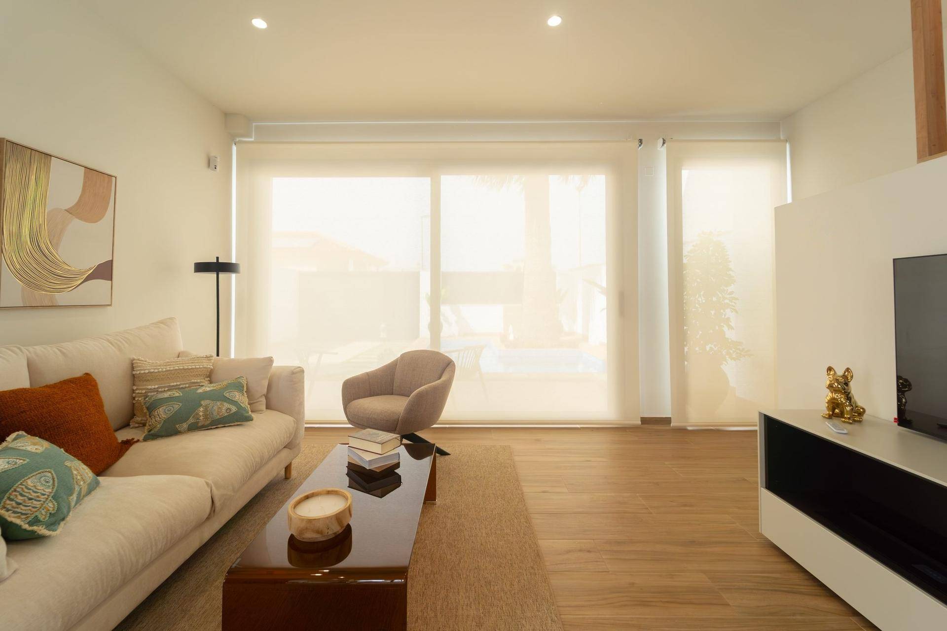 Helles Wohnzimmer mit beigem Sofa, Holzboden und großen Fenstern mit Rollos.