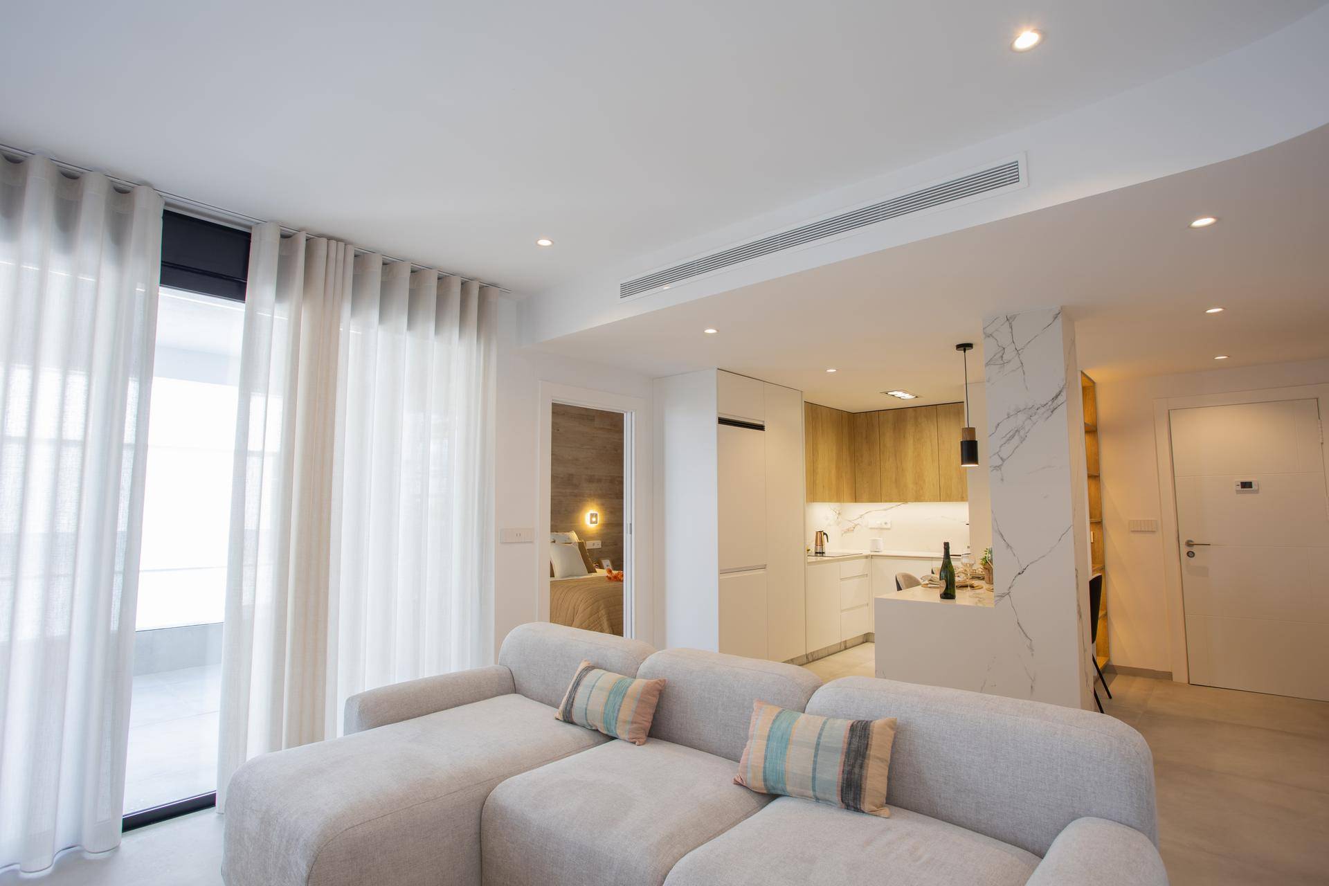 Offener Wohnbereich mit beigem Sofa, integrierter Küche und Klimaanlage in der Decke.