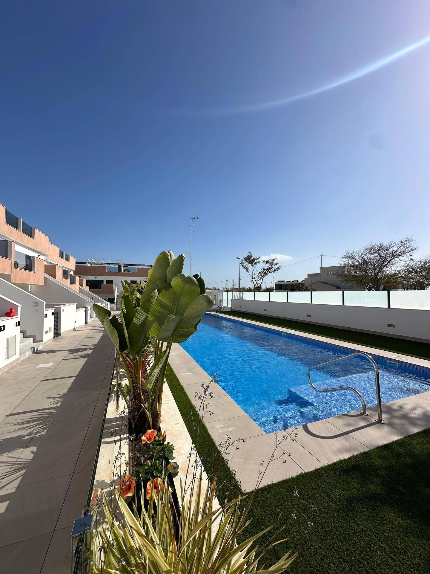 Sonniger Gehweg und Poolbereich mit tropischen Pflanzen und modernen Gebäuden im Hintergrund.