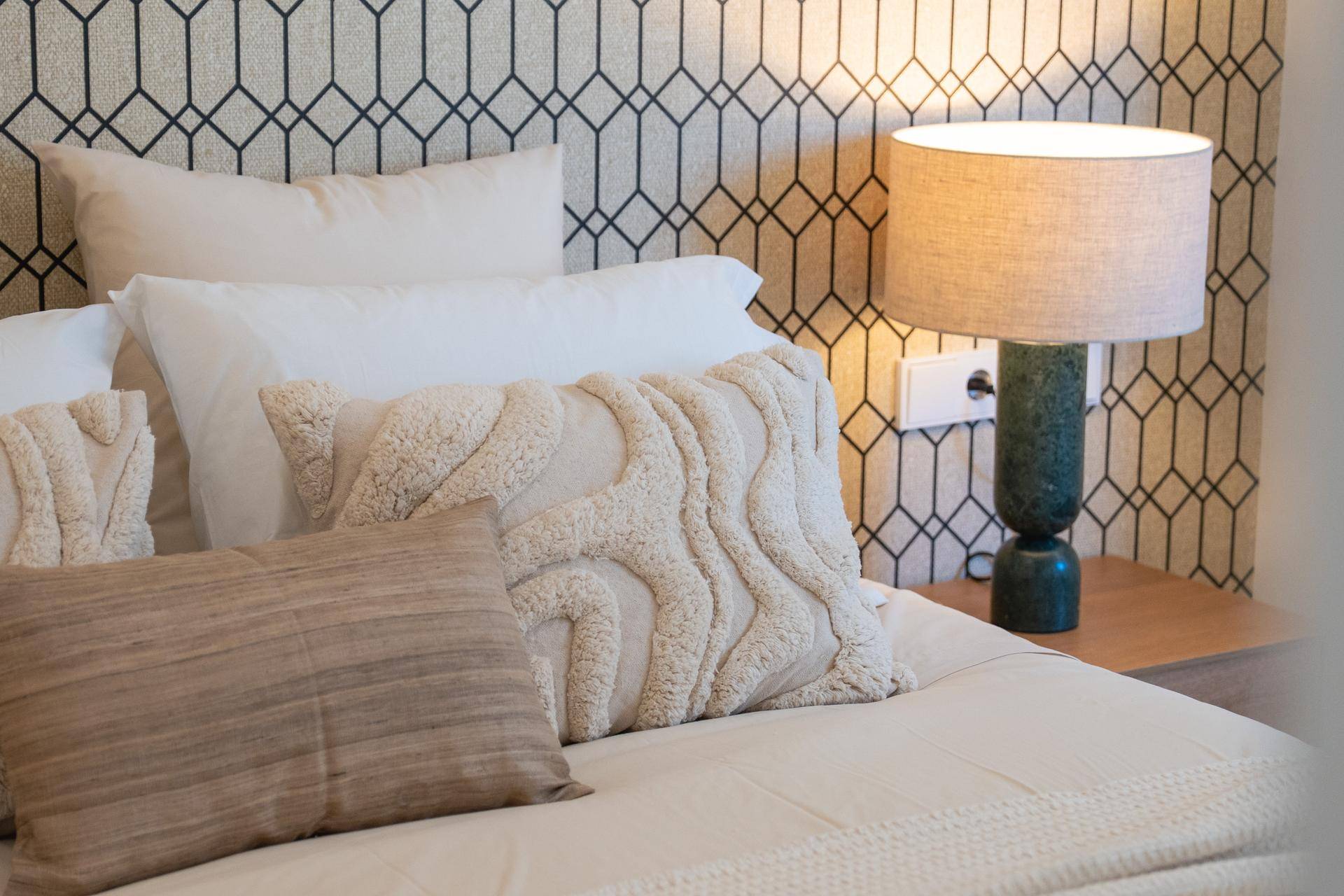 Gemütliches Bett mit dekorativen Kissen und einer eleganten Lampe vor einer gemusterten Wand.