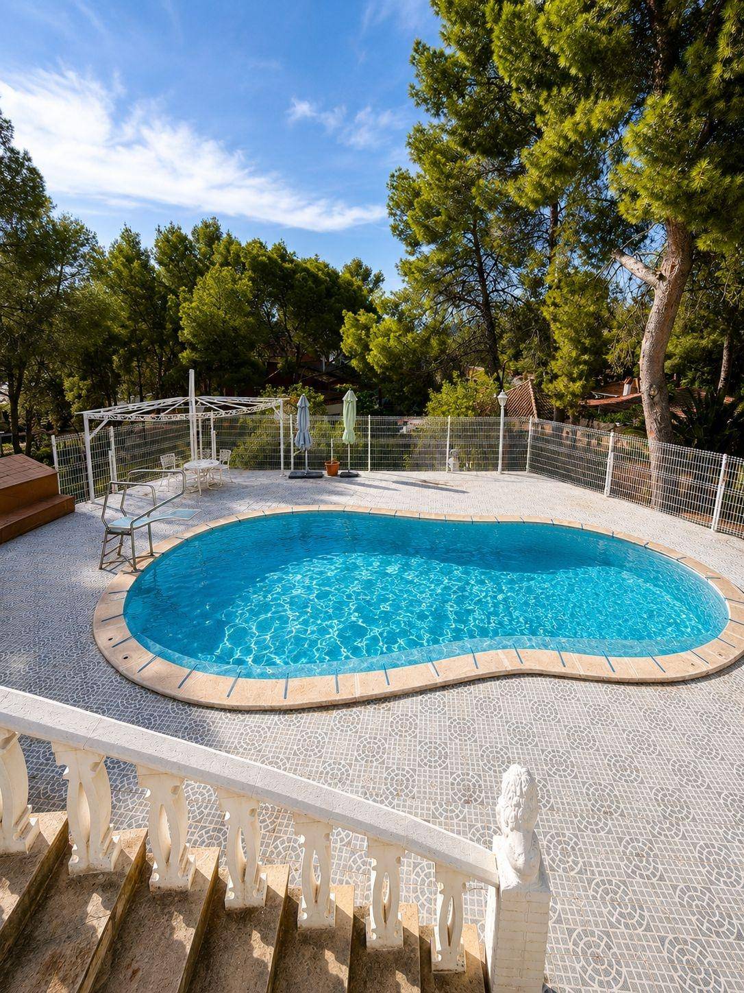 Nierenförmiger Poolbereich mit umliegender Terrasse und Treppenzugang, umgeben von hohen Bäumen unter blauem Himmel.