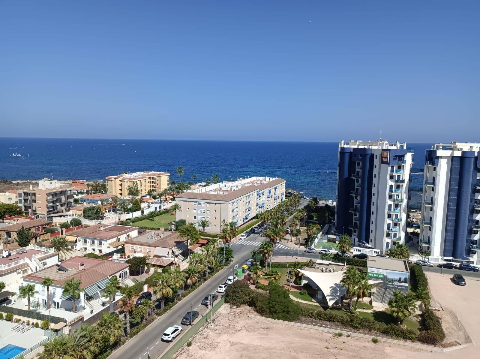 Panoramablick auf die Küstenlinie mit Gebäuden, Straße und dem weiten Meer unter blauem Himmel.