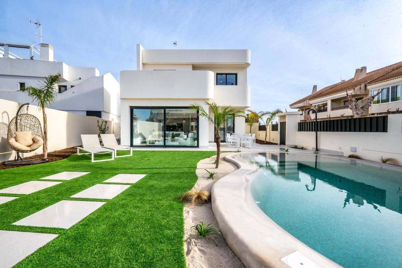 Moderner Gartenbereich mit Pool, Kunstrasen, Liegestühlen und einer Schaukel vor der weißen Villa.