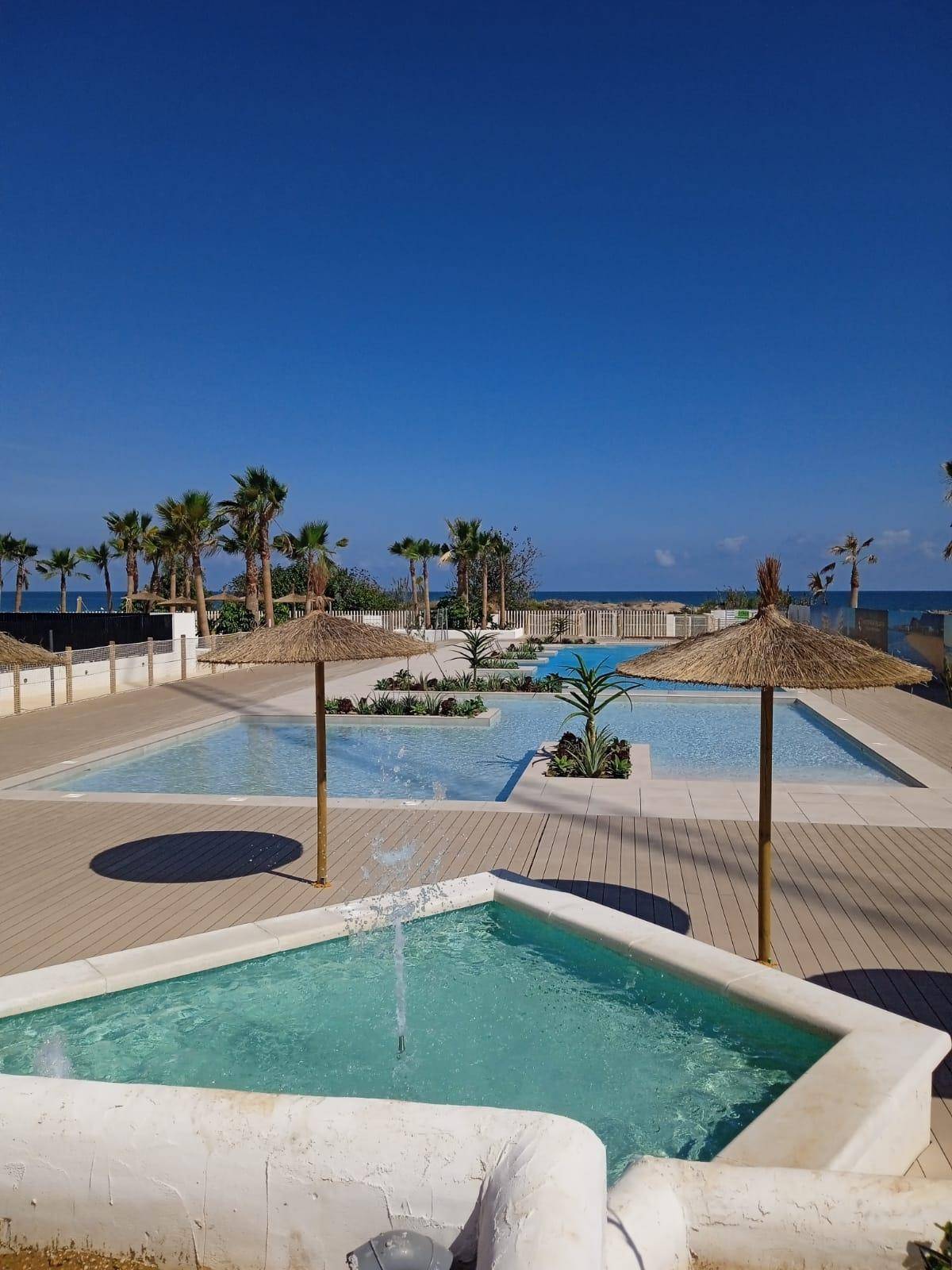 Detailansicht eines Brunnens im Poolbereich mit weiteren Pools, Palmen und Meerblick im Hintergrund.