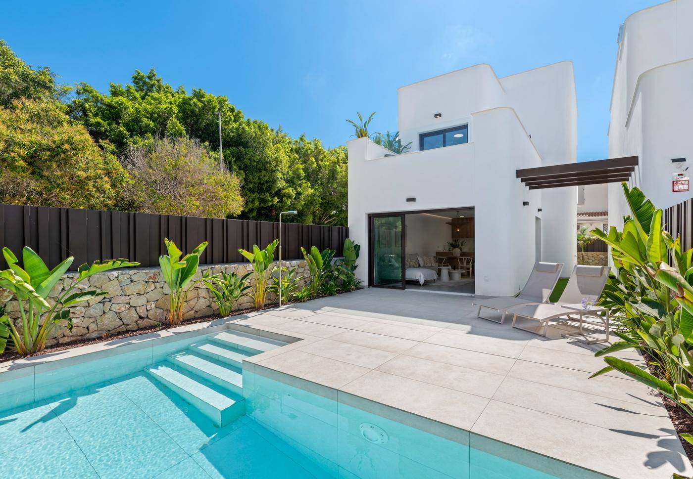 Sonniger Außenbereich mit privatem Pool, Liegestühlen und üppiger Bepflanzung hinter der modernen Villa.