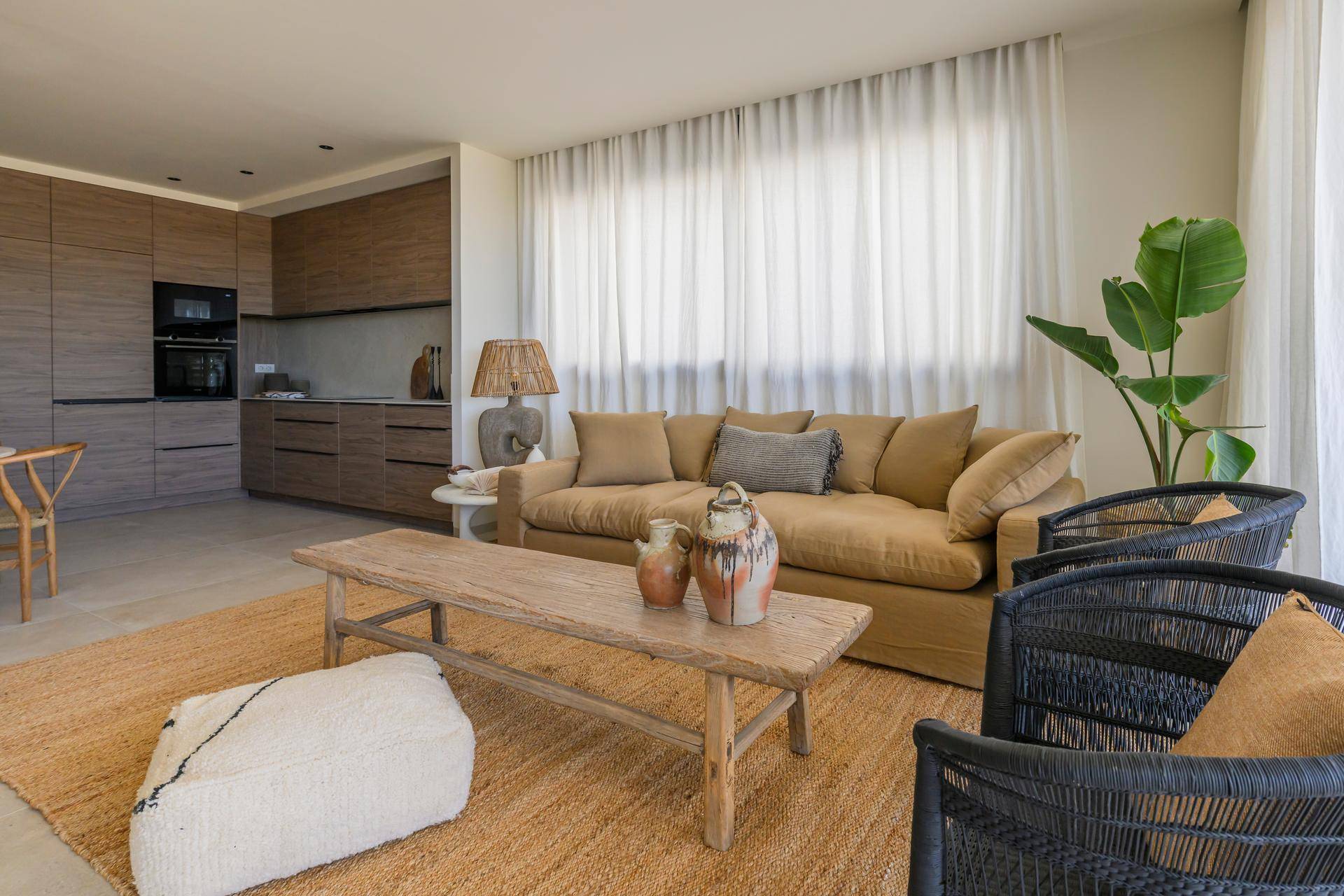 Heller Wohnbereich mit beigem Sofa, Holztisch und zwei schwarzen Korbstühlen, umgeben von transparenten Vorhängen.