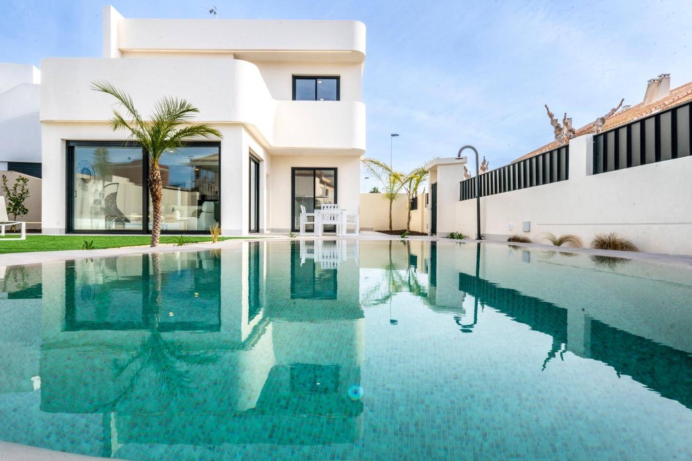 Großer Poolbereich mit reflektierendem Wasser, umgeben von einer modernen weißen Villa und Palmen.