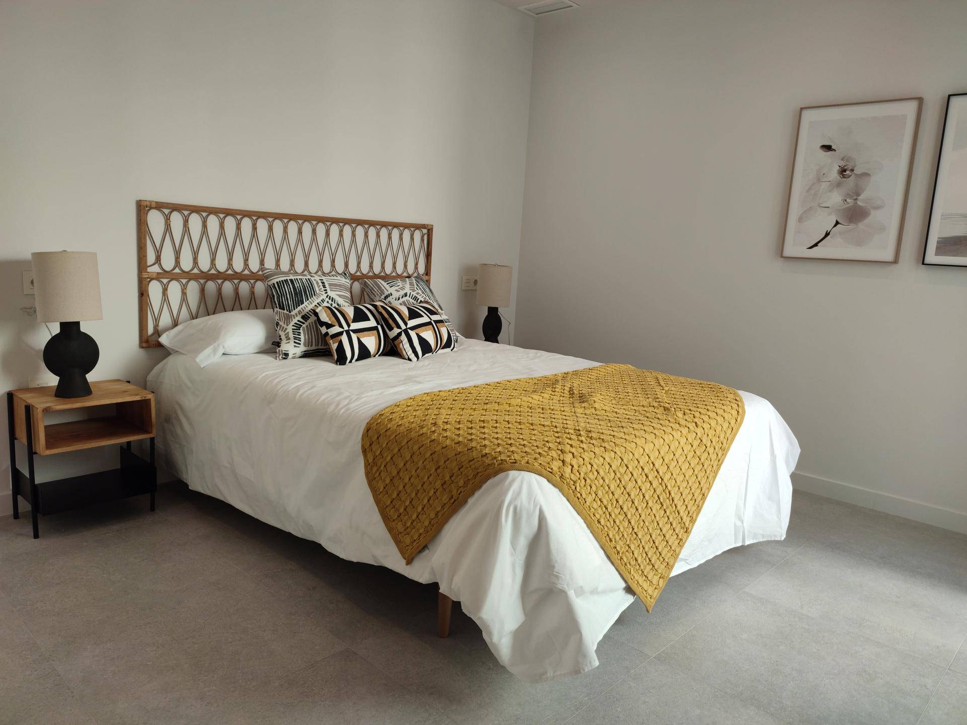 Helles Schlafzimmer mit einem Bett, Rattan-Kopfteil und einer gelben Tagesdecke in schlichter Gestaltung.