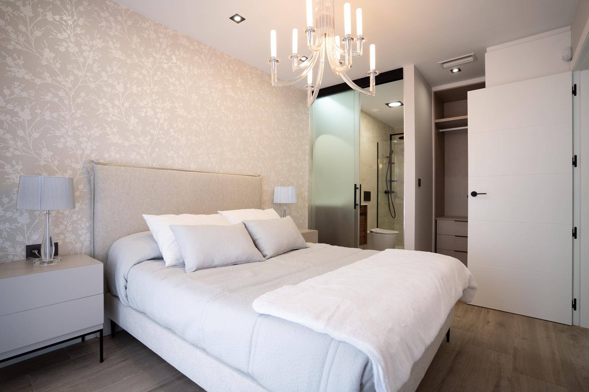 Schlafzimmer mit Doppelbett, floraler Tapete und integriertem Bad mit Glasschiebetür.